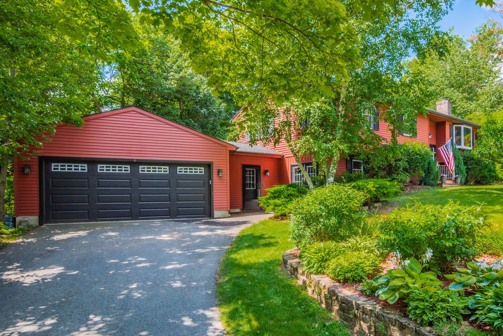 Red house with attached garage, surrounded by green trees and grass, on a sunny day.