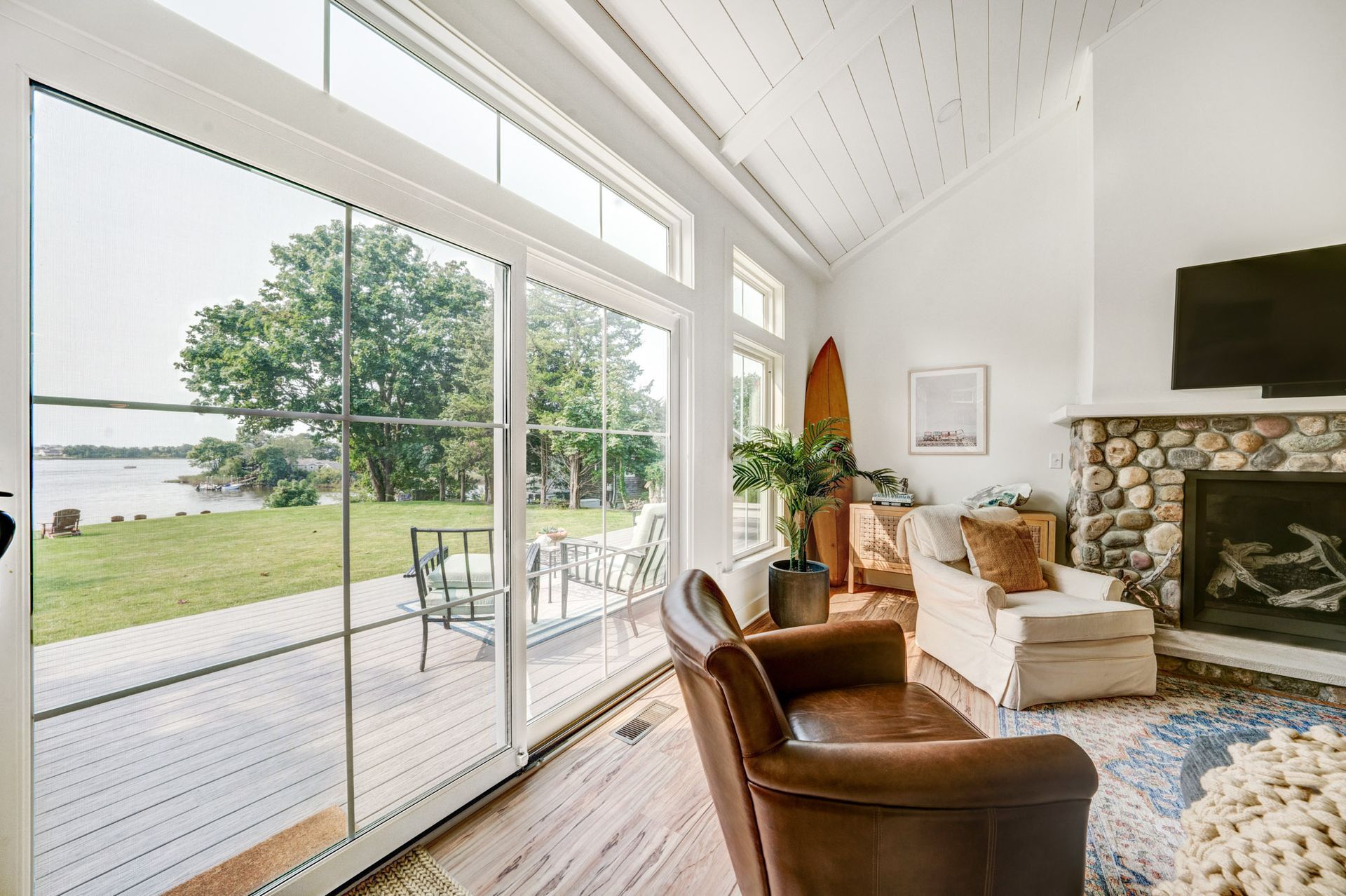 Living room with large windows overlooking a lake, stone fireplace, brown leather chair, and wooden surfboard.