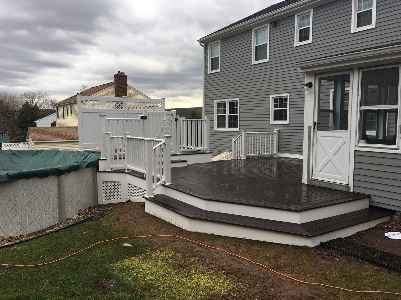 Gray house with a dark brown deck, white railing, and an above-ground pool under a cloudy sky.