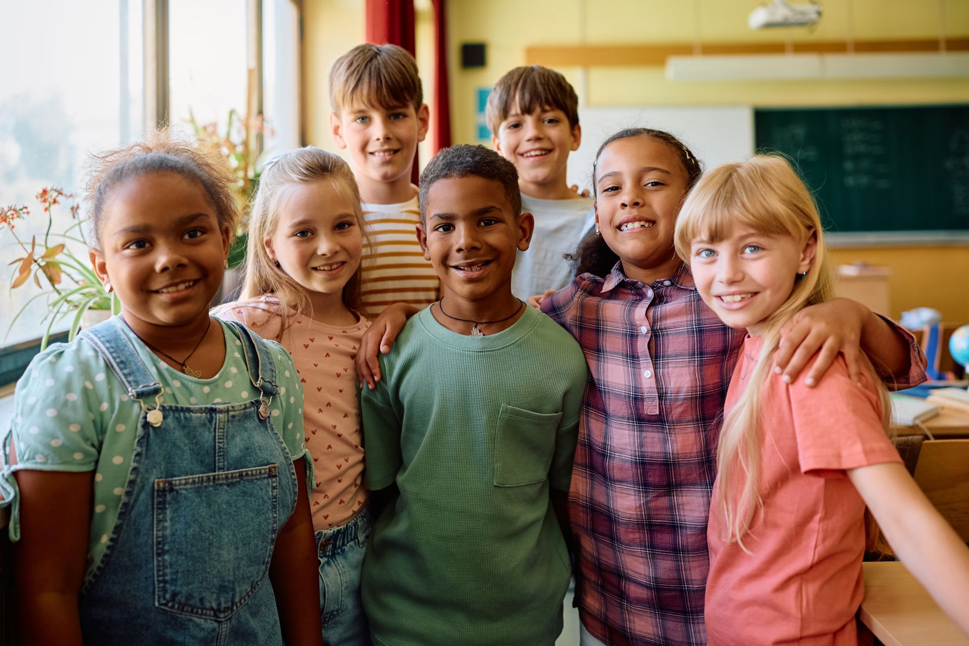 Group of children smiling in a classroom with a window, blackboard, and desk.