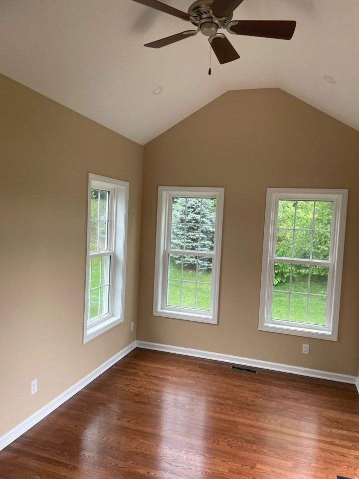 A room with brown wooden floor, tan walls, and three windows with white frames. A ceiling fan hangs above.