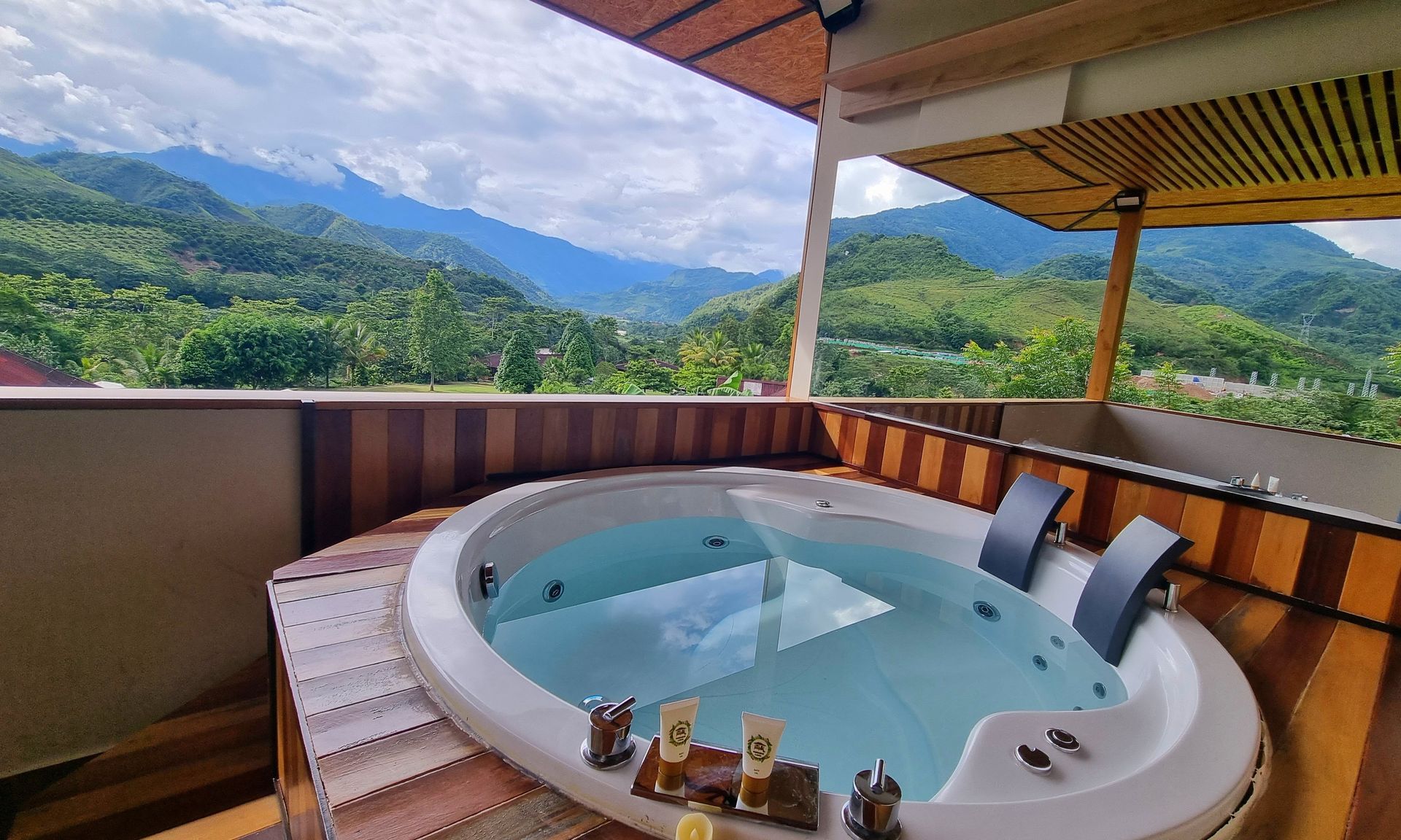 Bañera de hidromasaje al aire libre en un balcón de madera con vistas a las verdes montañas y a un cielo nublado.