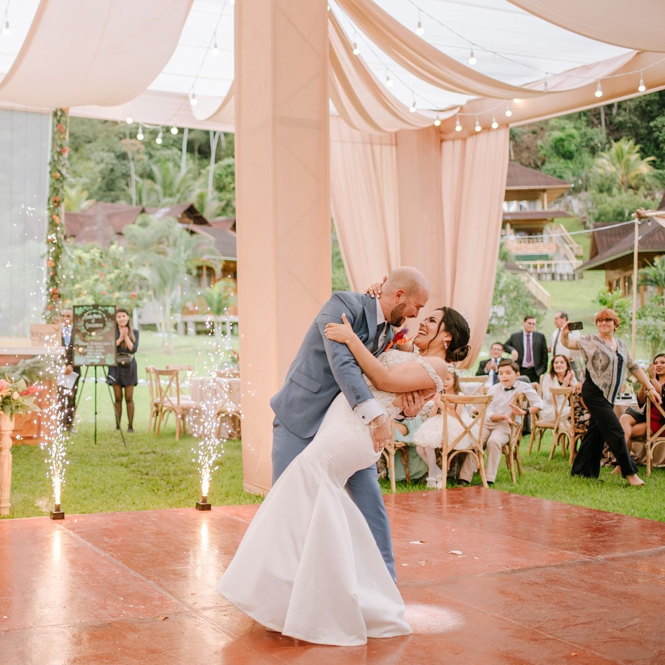Novios bailando en una recepción de boda al aire libre bajo cortinas blancas y decoración color melocotón.