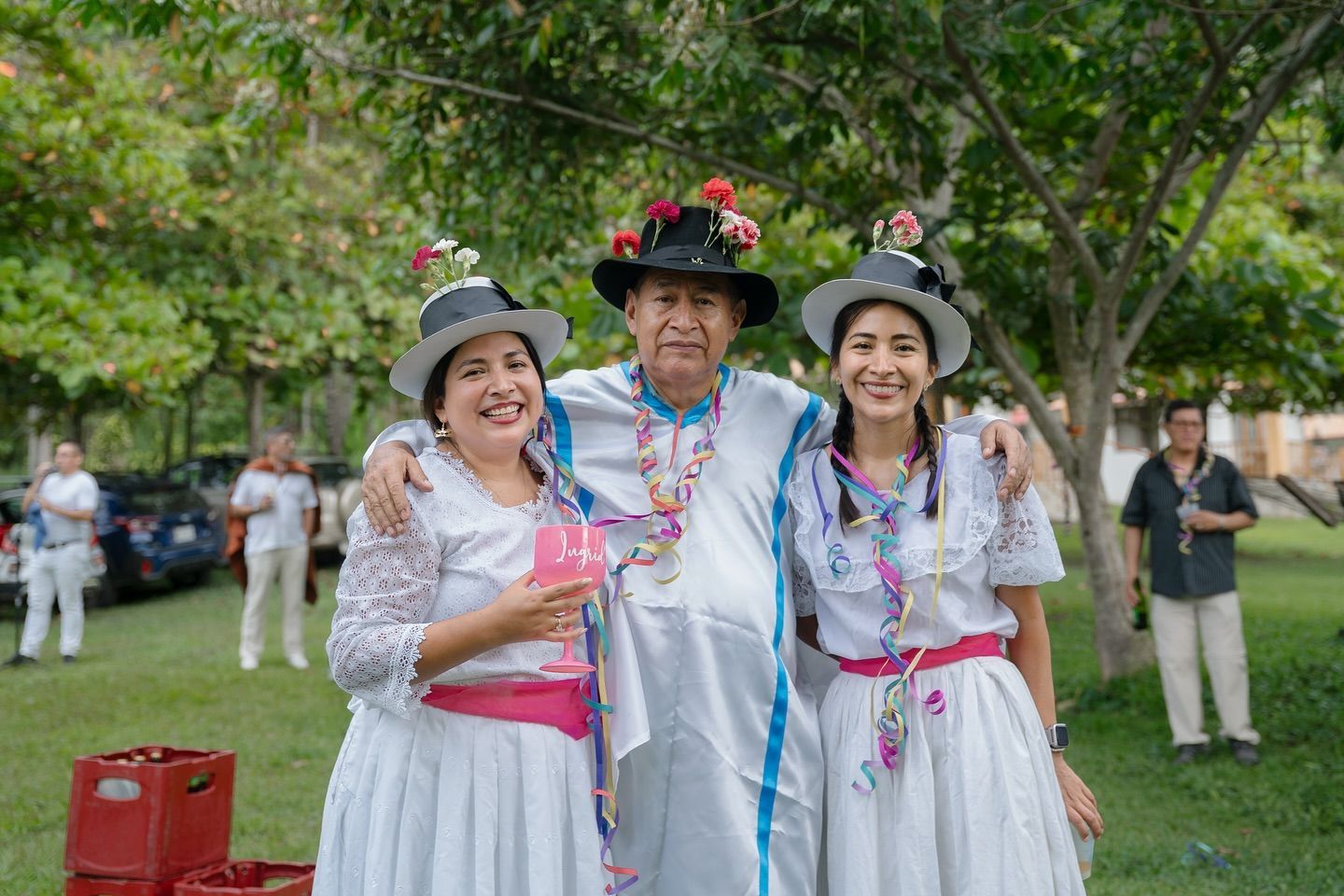 Tres personas sonrientes con sombreros festivos y collares de flores posando al aire libre en un parque.