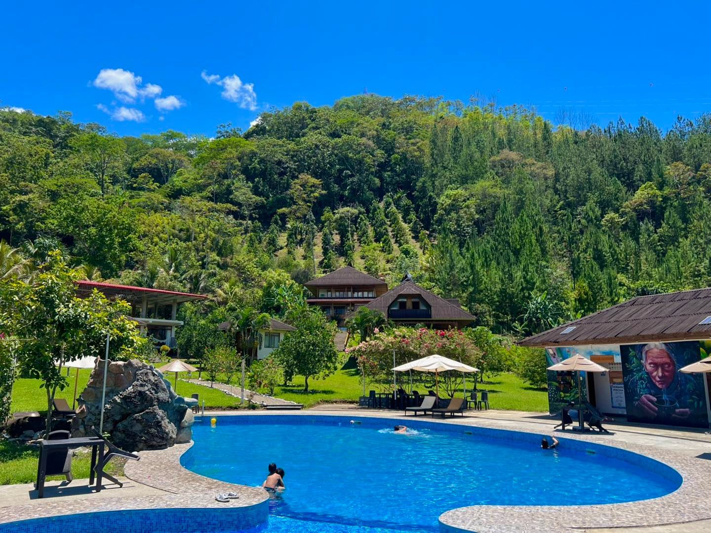 Piscina del complejo turístico con gente descansando, cabaña y una exuberante ladera verde bajo un cielo azul.