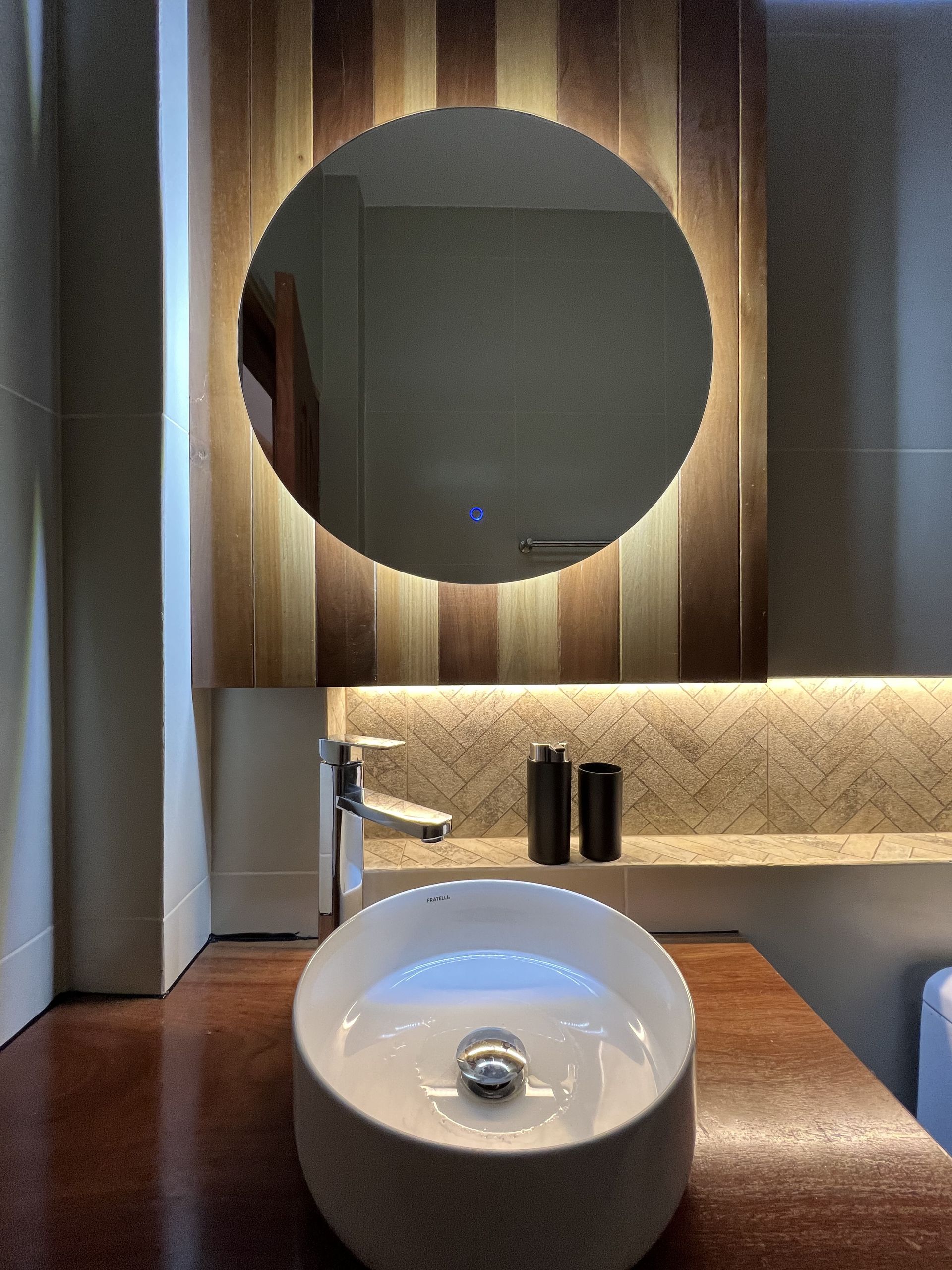 Lavabo moderno con espejo redondo, revestimiento de madera e iluminación ambiental suave.