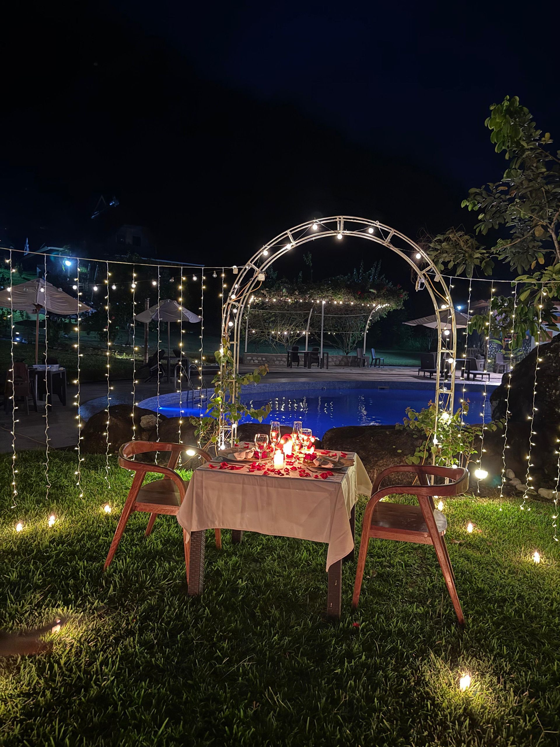 Cena romantica al aire libre con mesa iluminada con velas, guirnaldas de luces y un arco iluminado junto a la piscina.