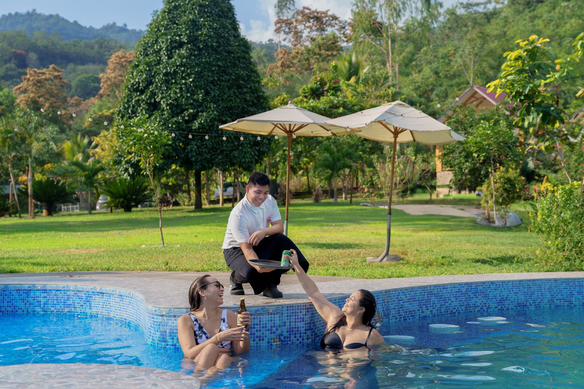 En una piscina, varias personas alzan a un niño hacia un hombre sentado al borde de la misma, en un jardín tropical.