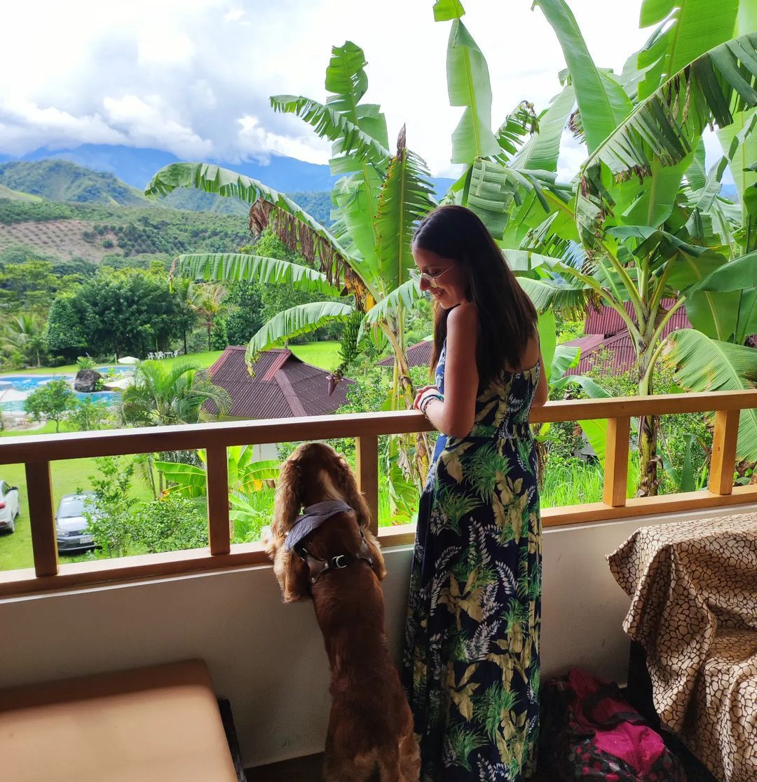 Mujer y perro en un balcón con vistas a exuberantes montañas verdes y plataneros.