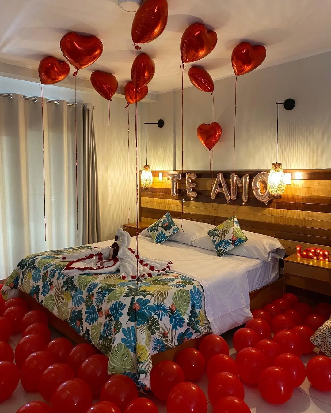 Habitación romántica con globos rojos, decoración en forma de corazón y dos camas decoradas bajo luces cálidas.