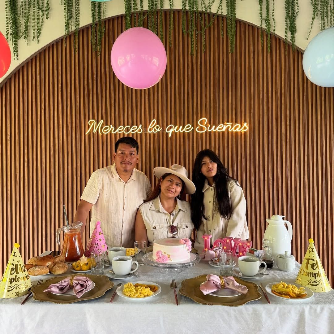 Tres personas posando en una mesa de cumpleaños con un globo rosa, pastel y gorros de fiesta, con un fondo de madera.