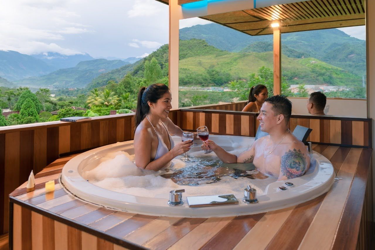 Una pareja brinda en un jacuzzi redondo al aire libre con vistas a la montaña y una terraza de madera al atardecer.