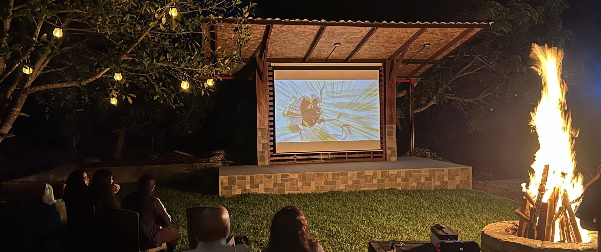 Proyección de cine al aire libre por la noche, con una fogata encendida junto a una película proyectada en un escenario de madera.