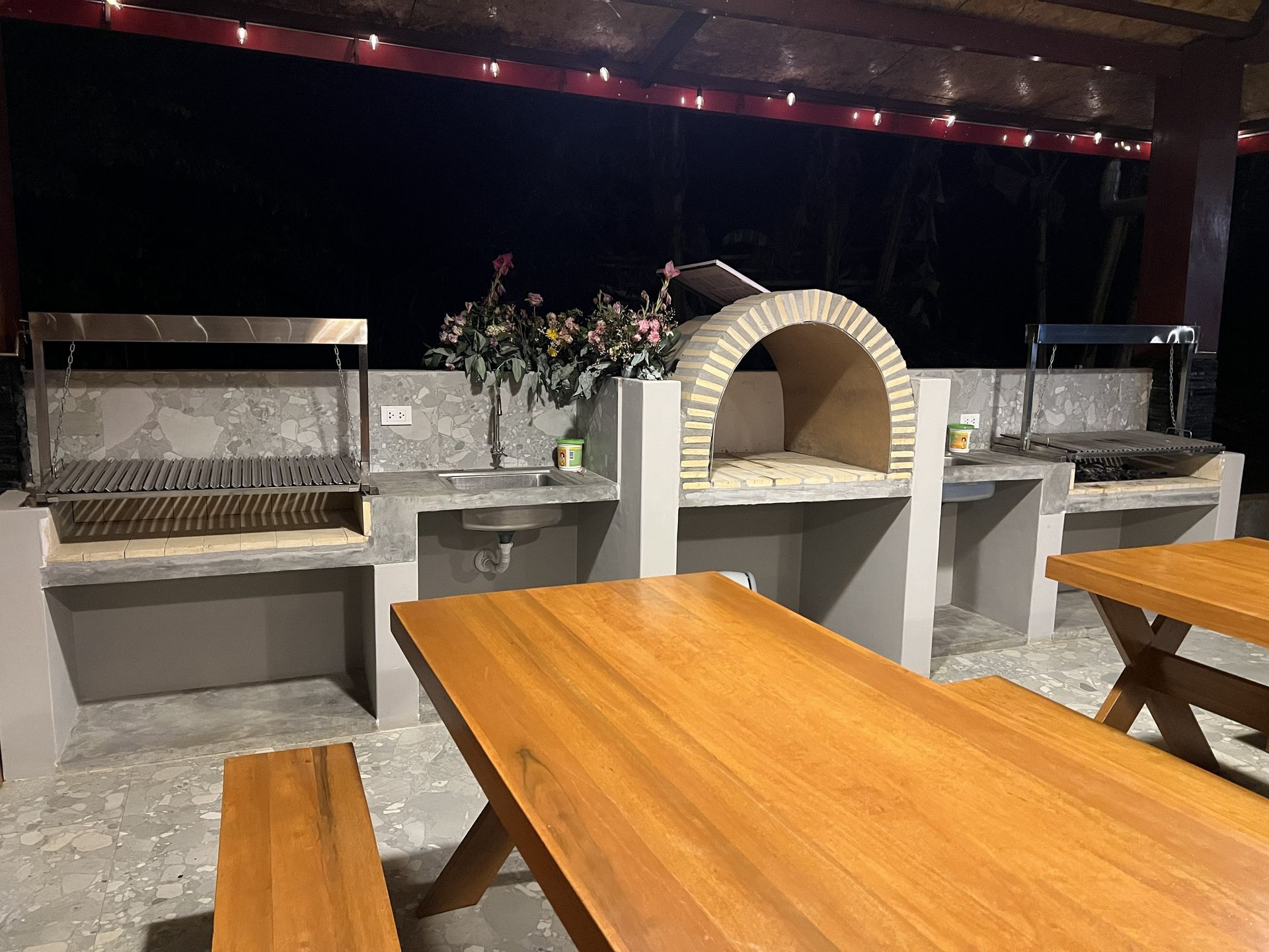 Patio exterior con parrillas de piedra, horno de pizza arqueado y mesas de picnic de madera por la noche.