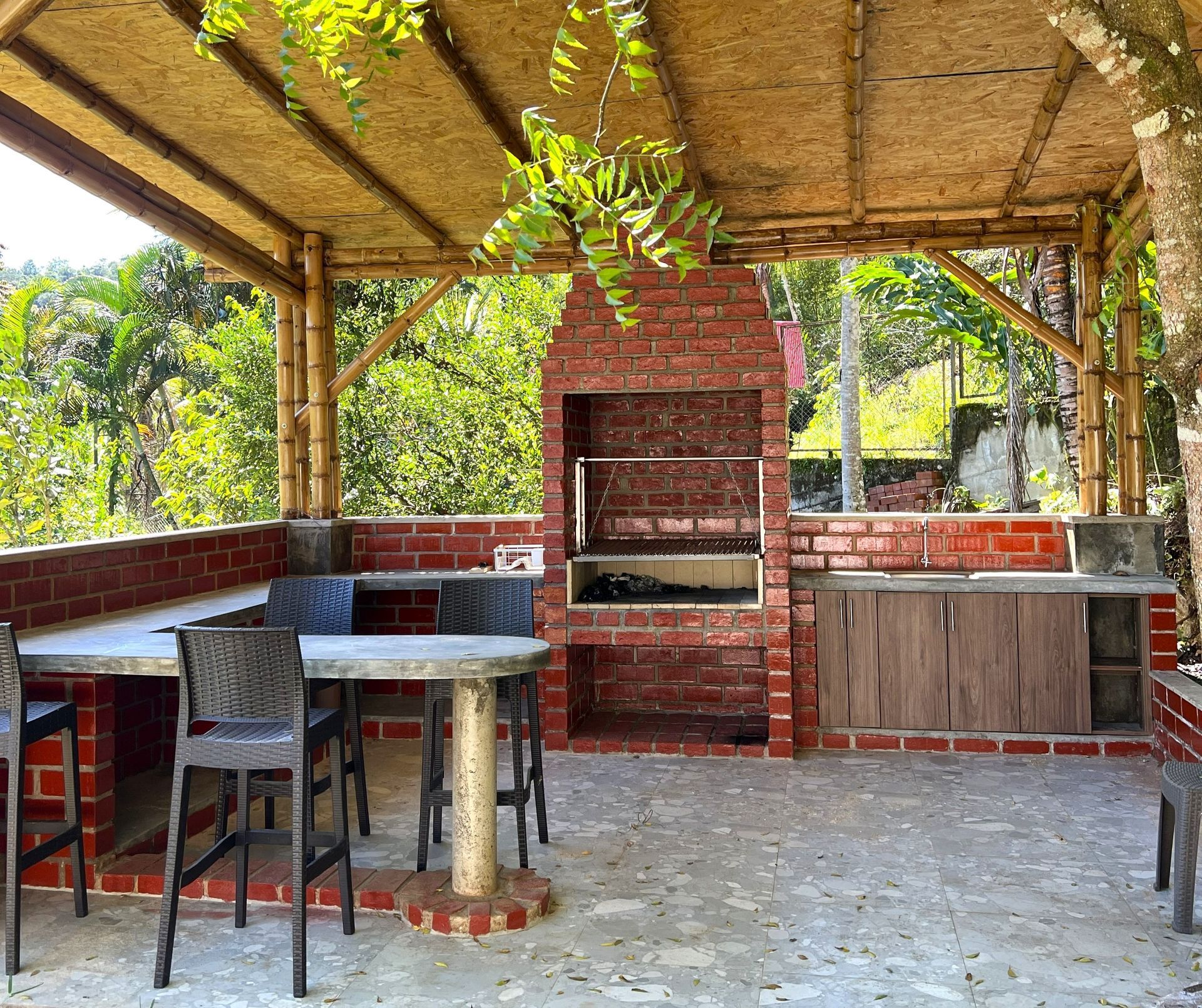 Zona de barbacoa al aire libre de ladrillo con techo de paja, mesas y taburetes en un entorno ajardinado.