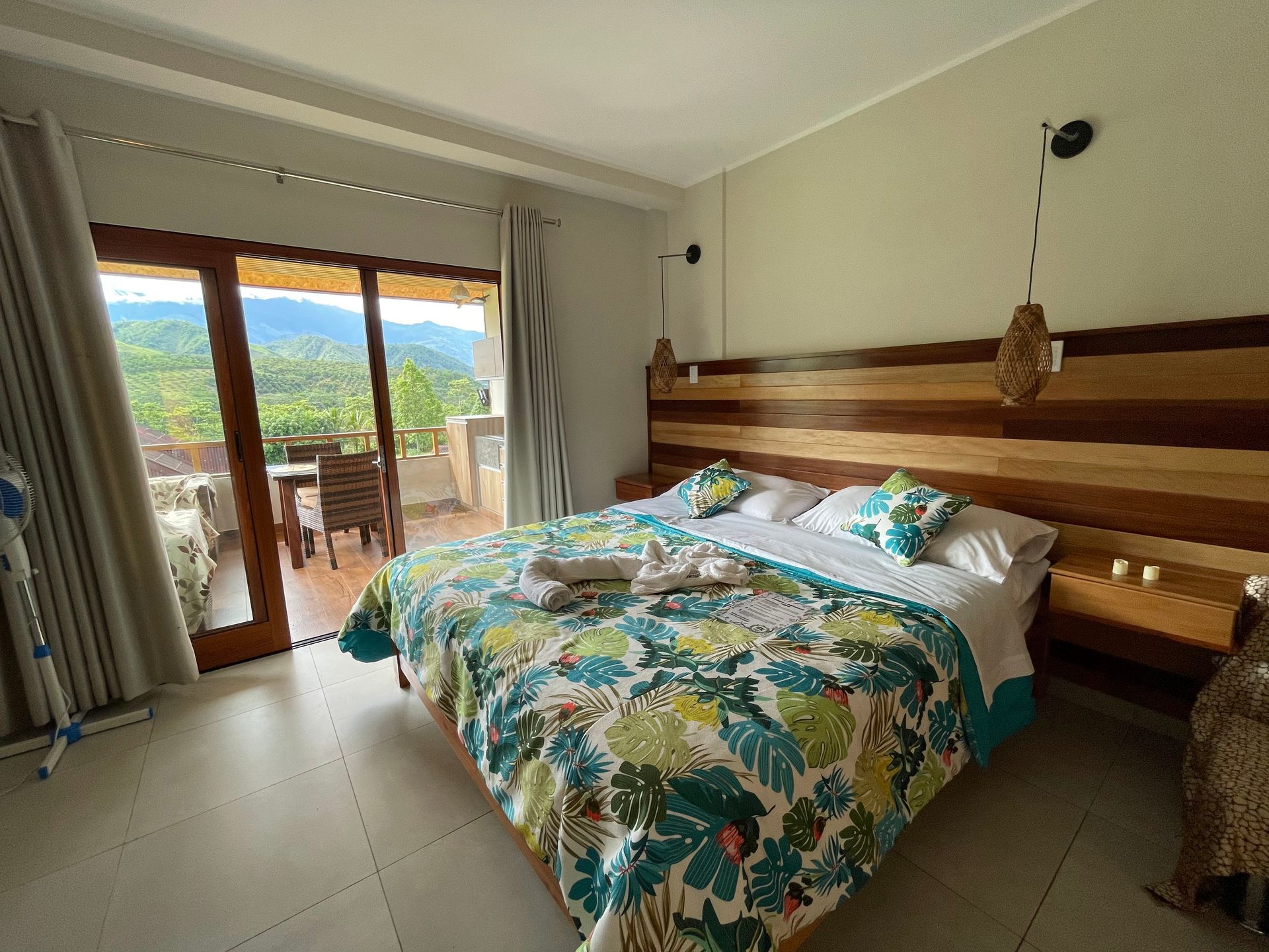 Habitación luminosa con colcha floral, cabecero de madera y puertas que dan a un balcón con vistas a la montaña.