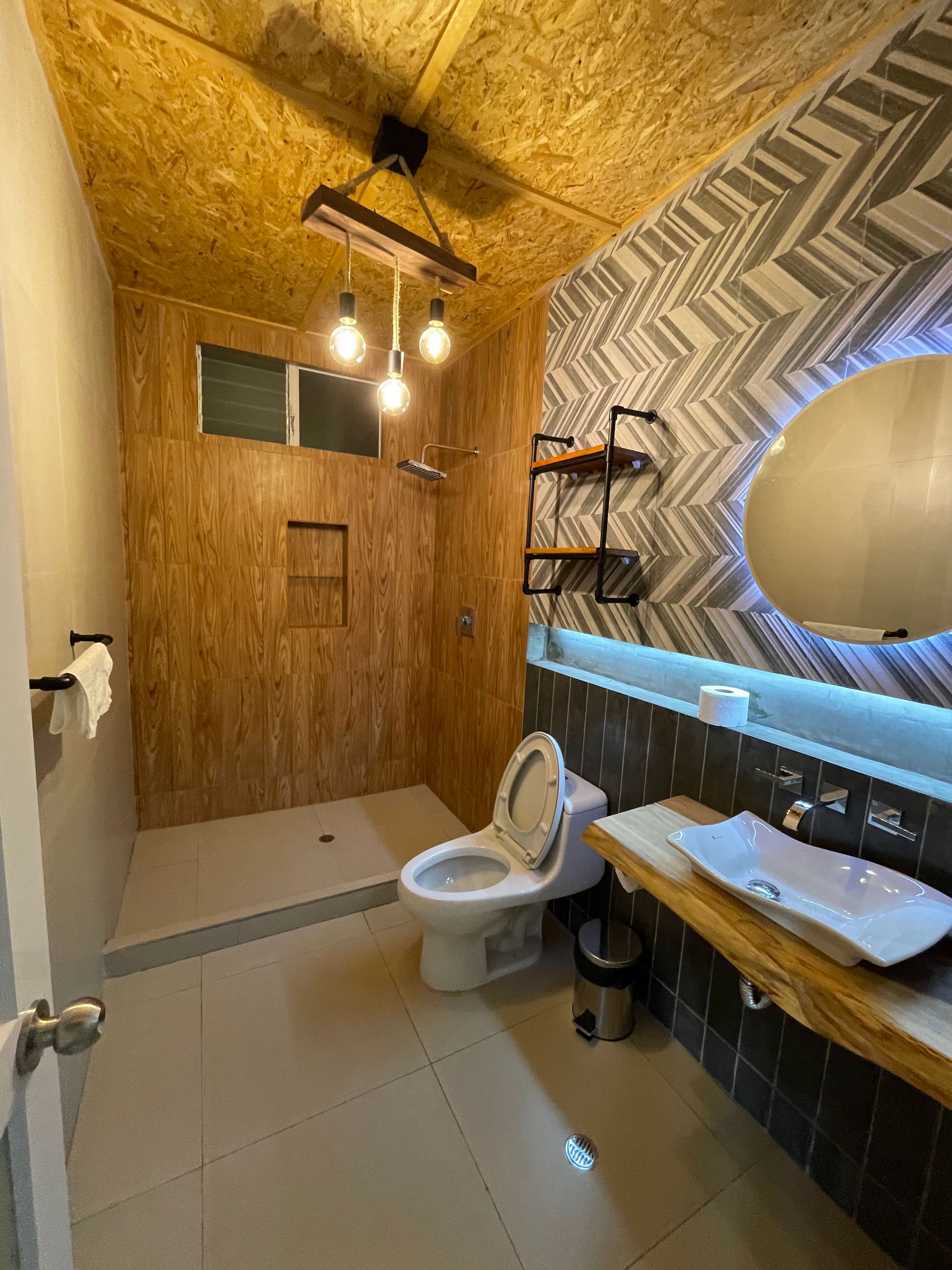 Pequeño baño rústico con paredes revestidas de madera, inodoro, lavabo, espejo y nicho para la ducha.