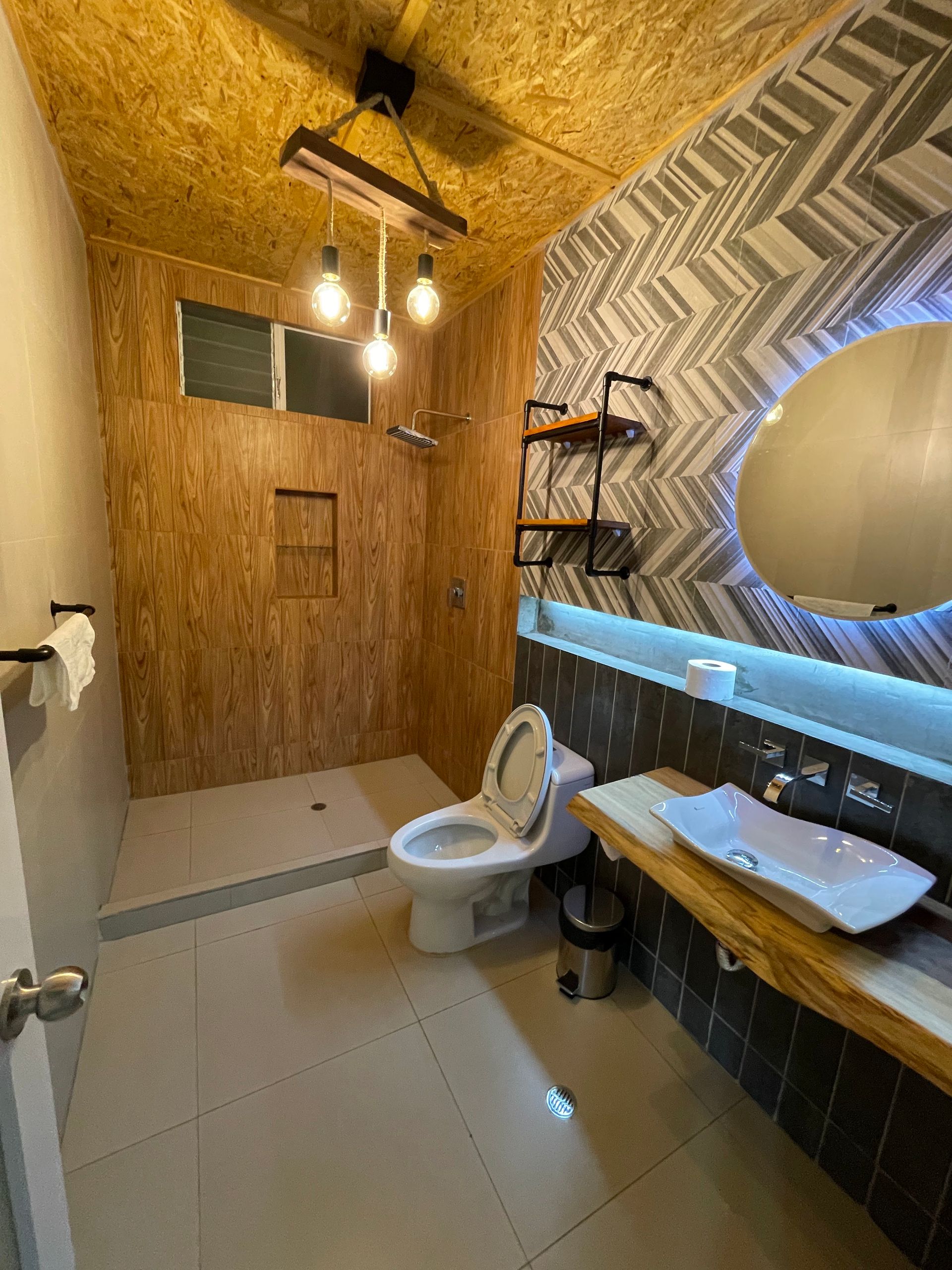 Baño moderno y compacto con paredes de madera, inodoro, lavabo, espejo redondo y suelo de baldosas.