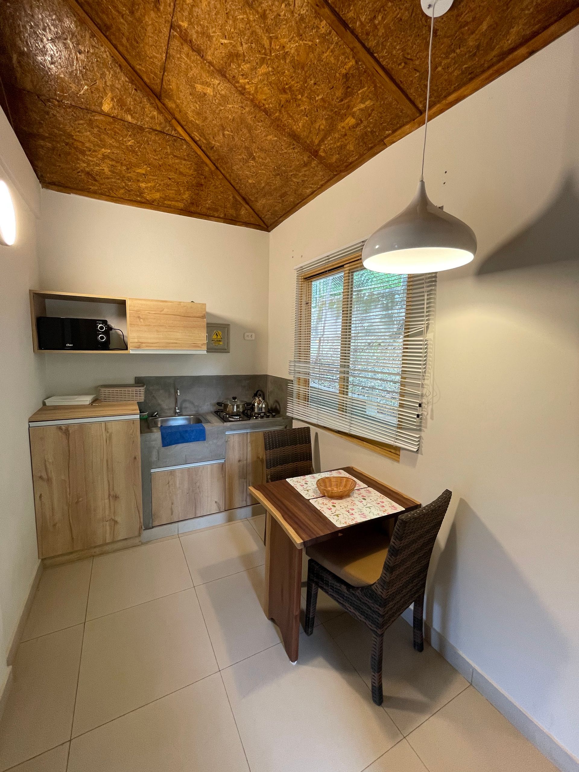 Pequeño rincón de cocina rústica con armarios de madera, mesa de mimbre, lámpara colgante y persianas.