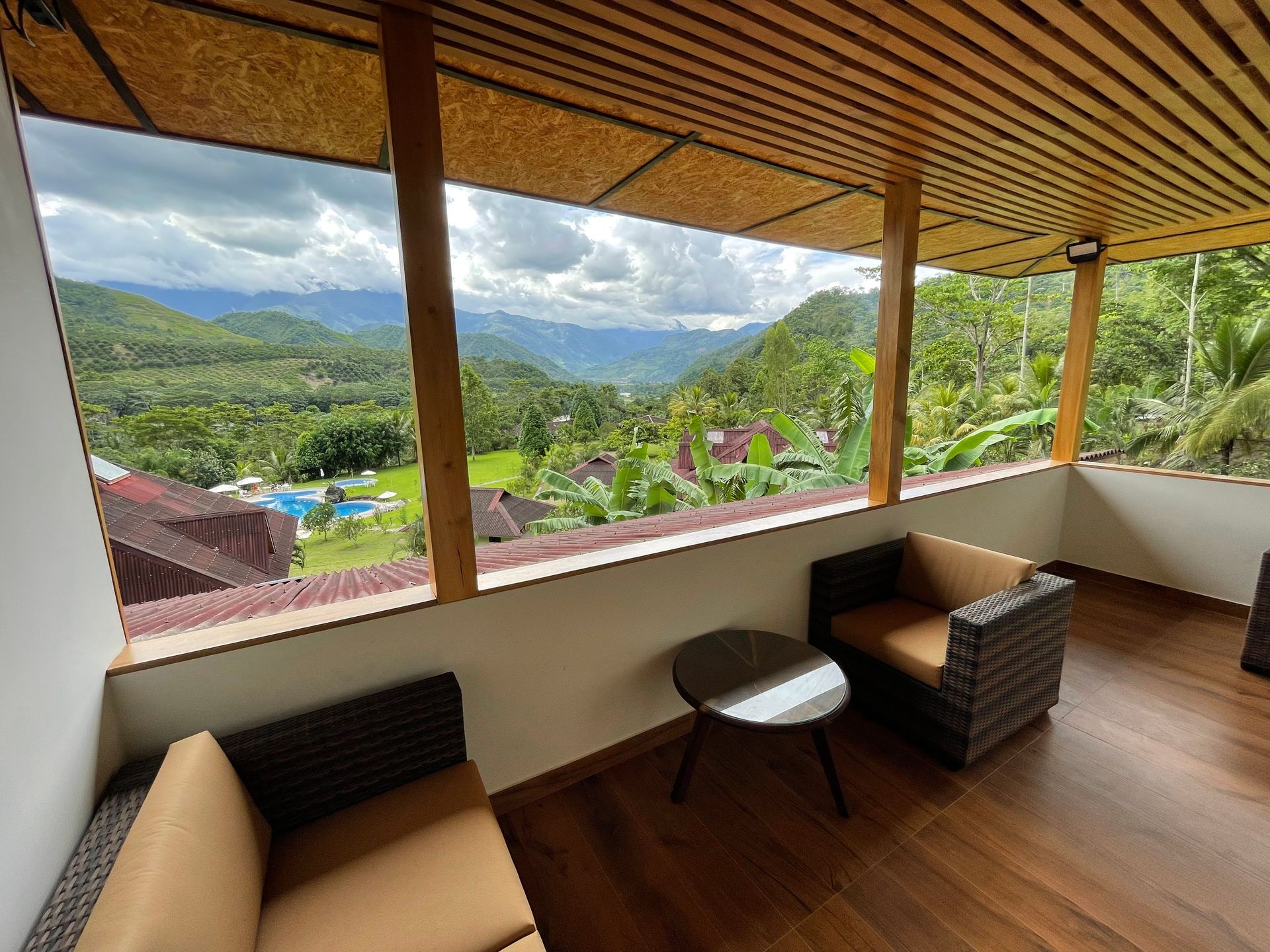 Balcón cubierto con sillas y mesa de mimbre con vistas a un exuberante valle de montaña verde.