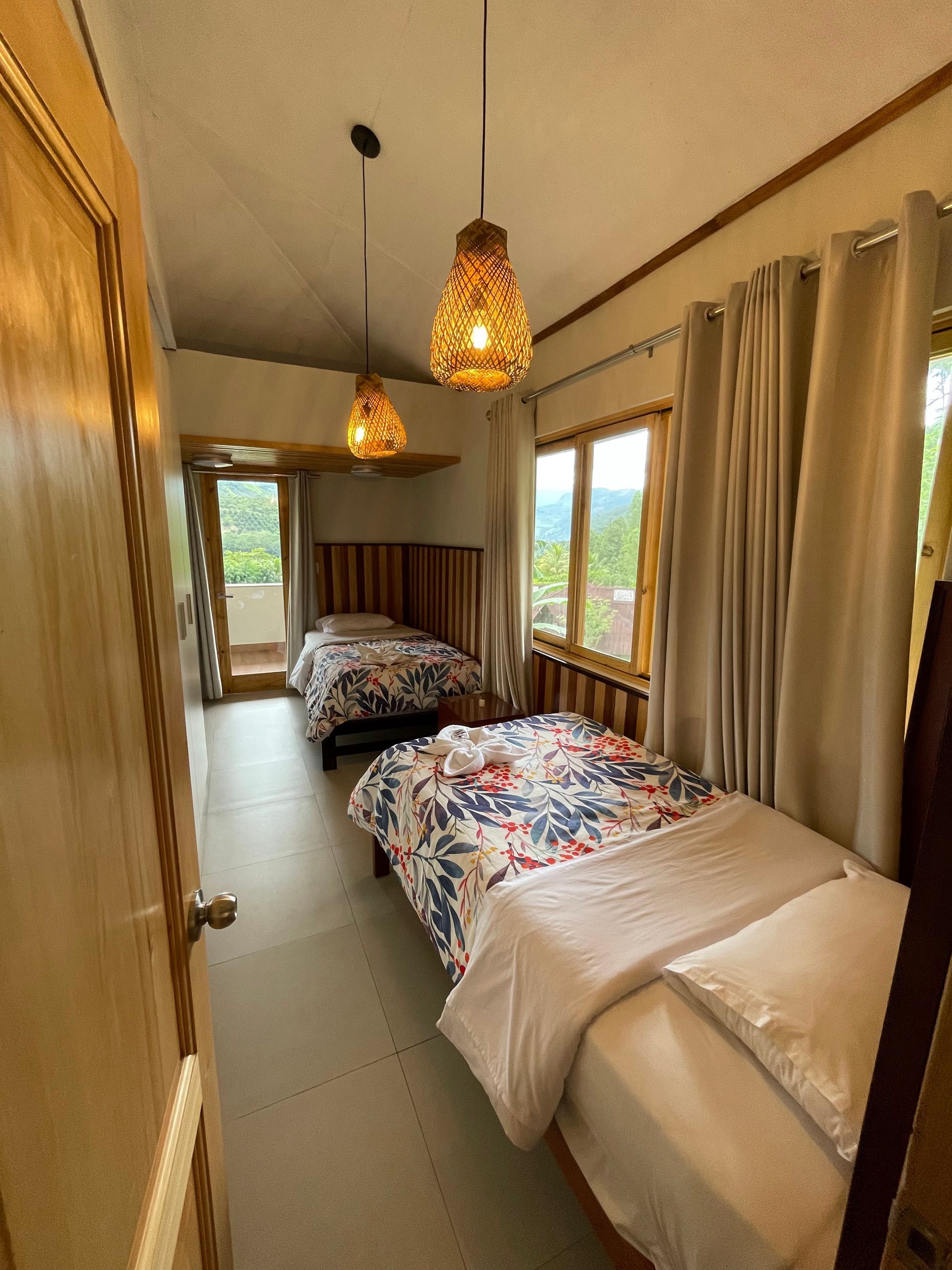 Habitación estrecha con dos camas, lámparas colgantes y grandes ventanas con cortinas beige.