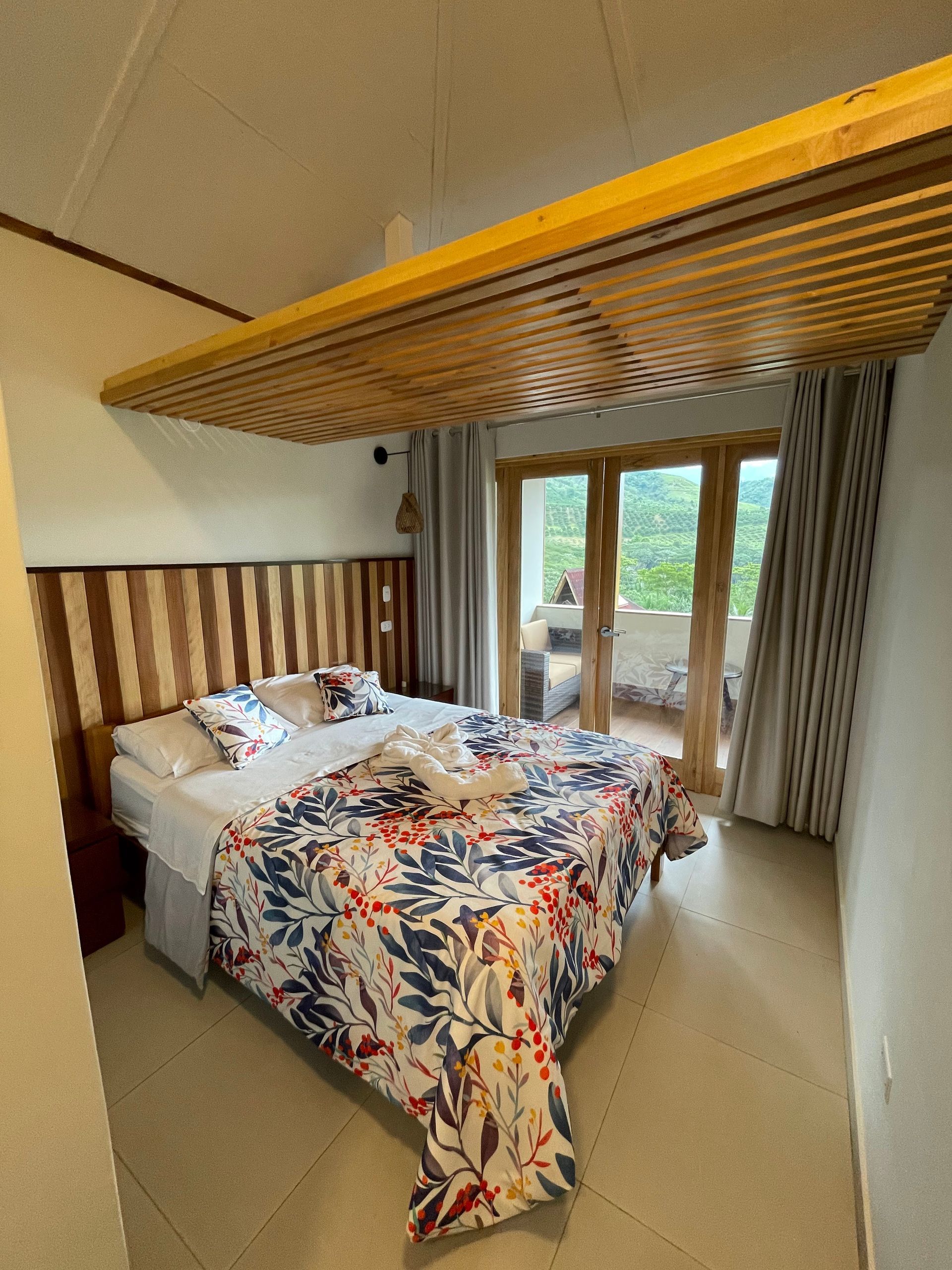 Dormitorio con colcha floral, cabecero de listones de madera y puertas correderas que dan a un balcón con vegetación exterior.