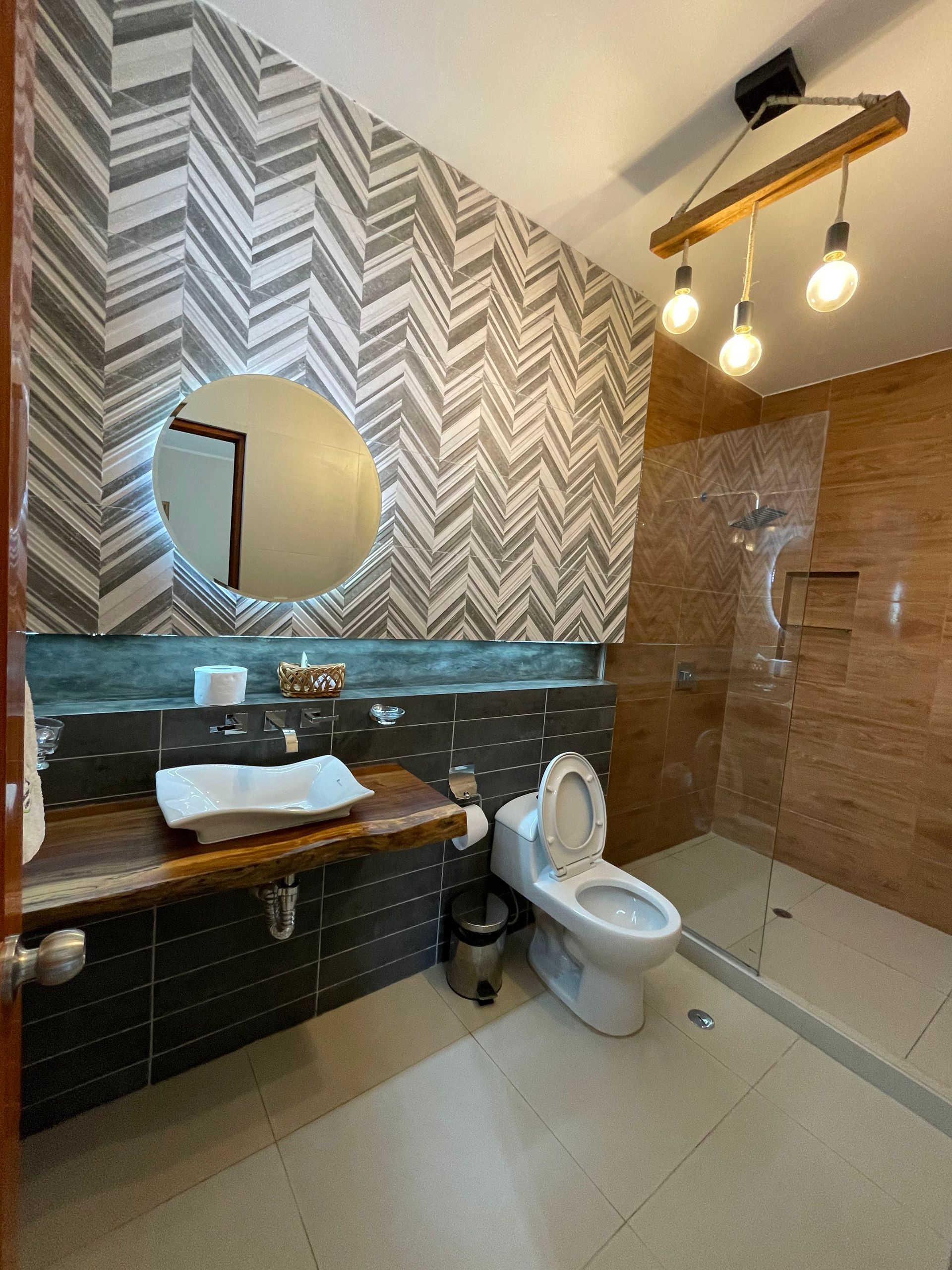 Baño moderno con azulejos estampados, espejo redondo, lavabo, inodoro y mampara de ducha de cristal.