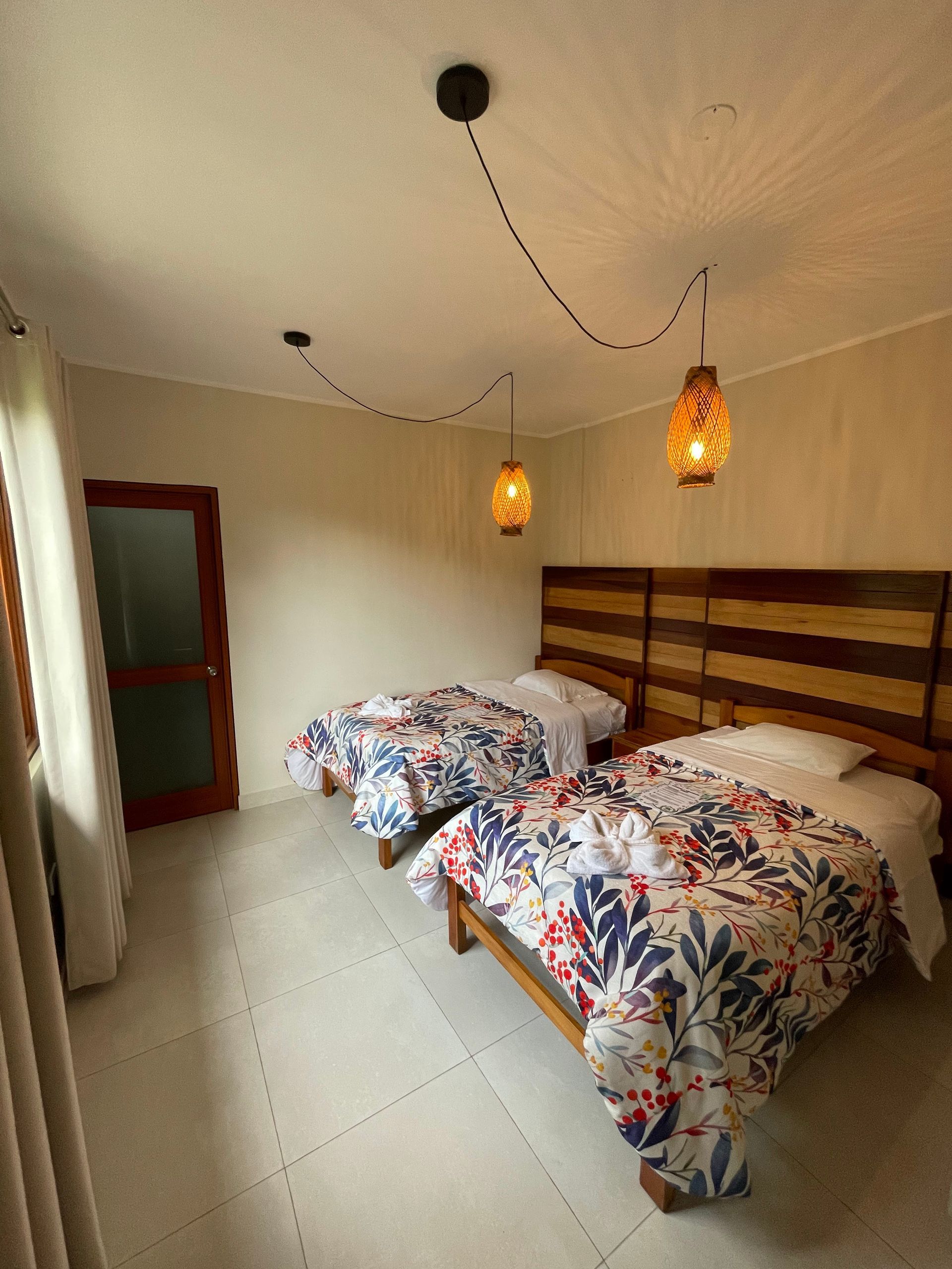 Habitación pequeña con dos camas individuales, colchas estampadas con flores, lámparas colgantes y cabecero de madera.