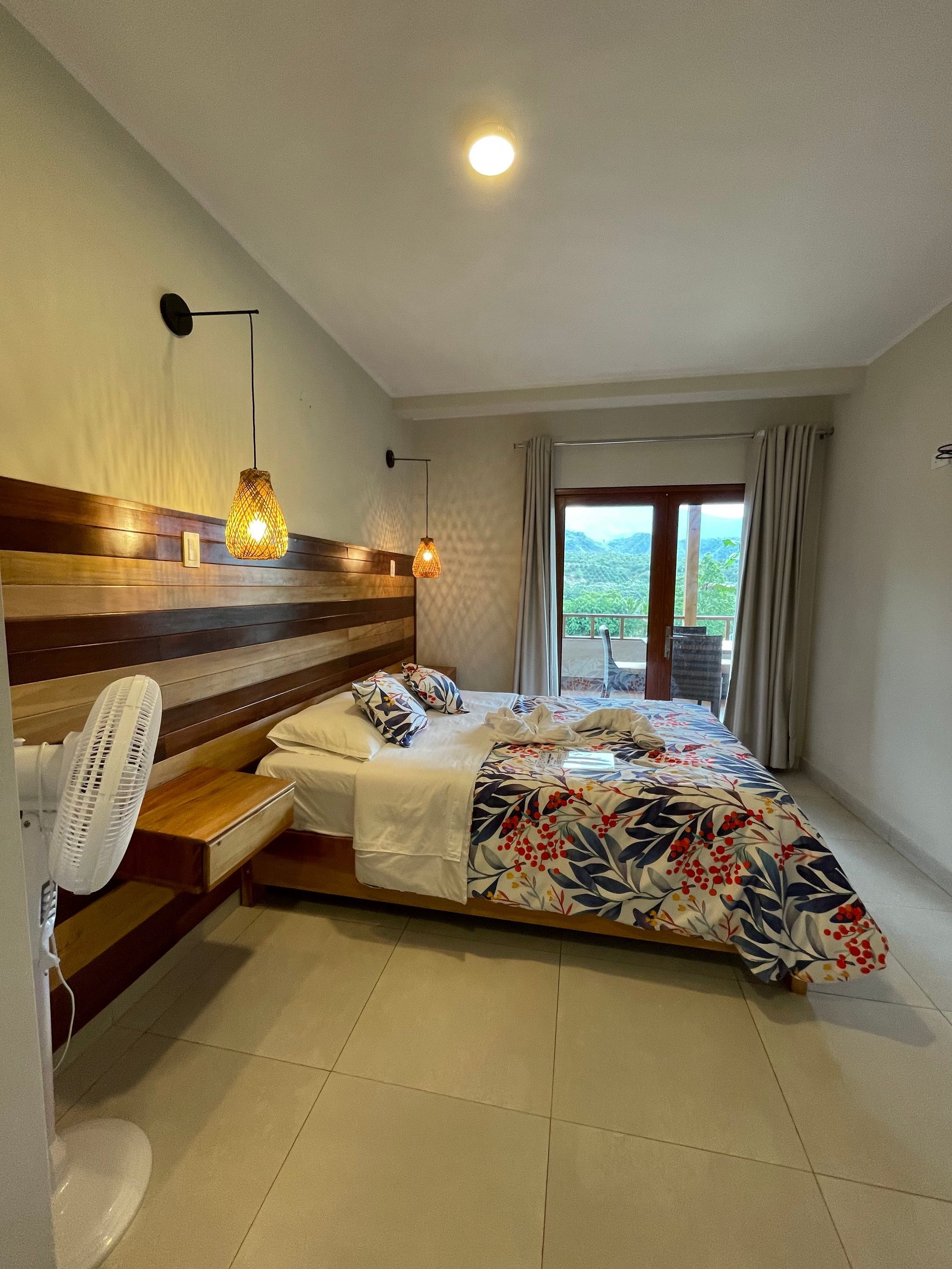 Acogedora habitación con cama, lámpara colgante, escritorio de madera y puertas que dan al balcón y dejan entrar la luz natural.