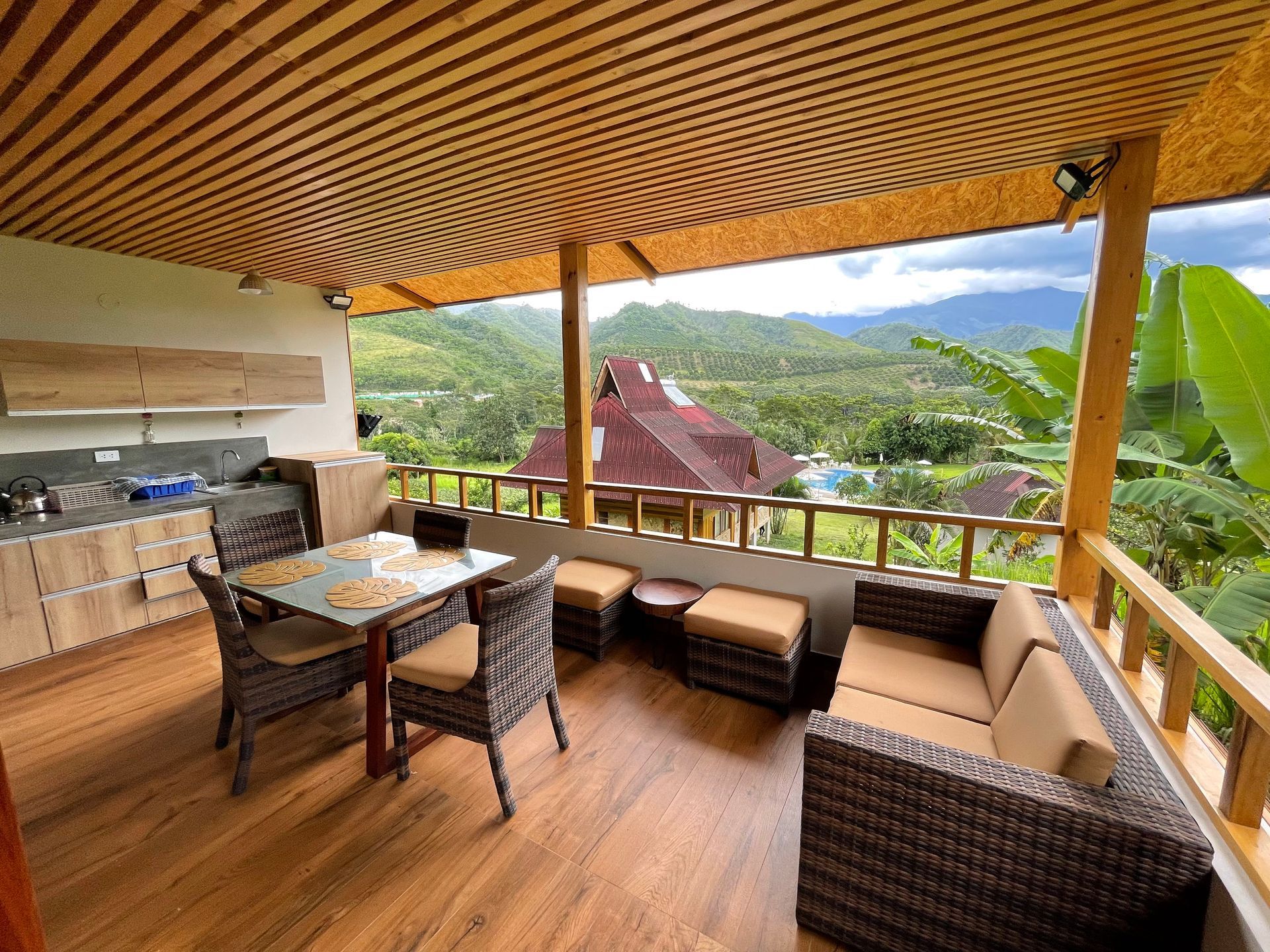 Terraza cubierta con asientos de mimbre, mesa de comedor y vistas al valle de la montaña bajo un techo de madera a rayas.