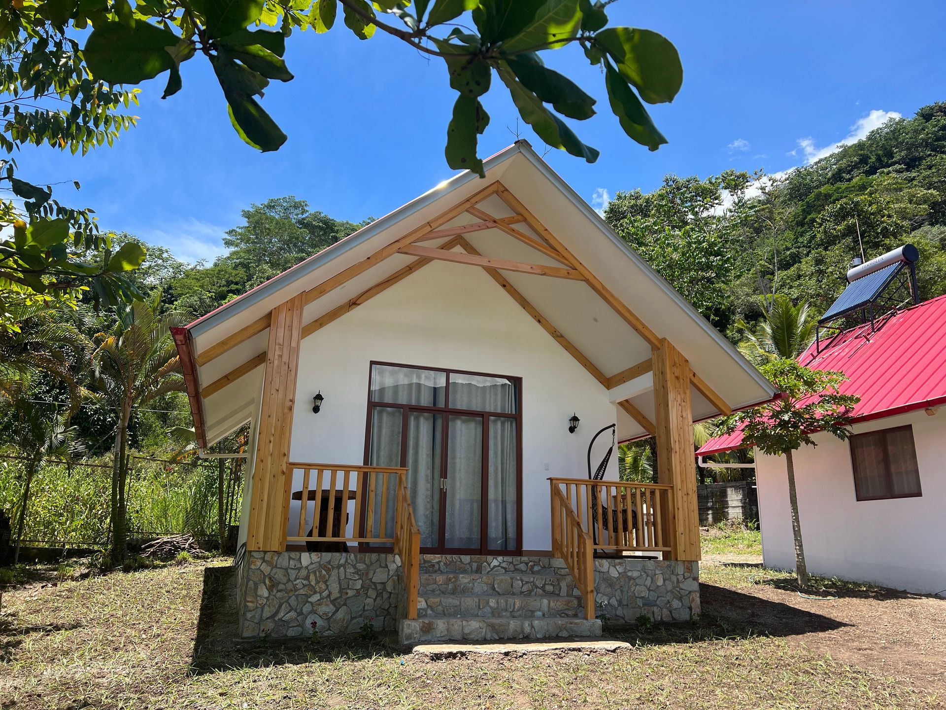 Pequeña casita blanca con porche de madera y techo rojo en un entorno tropical soleado.