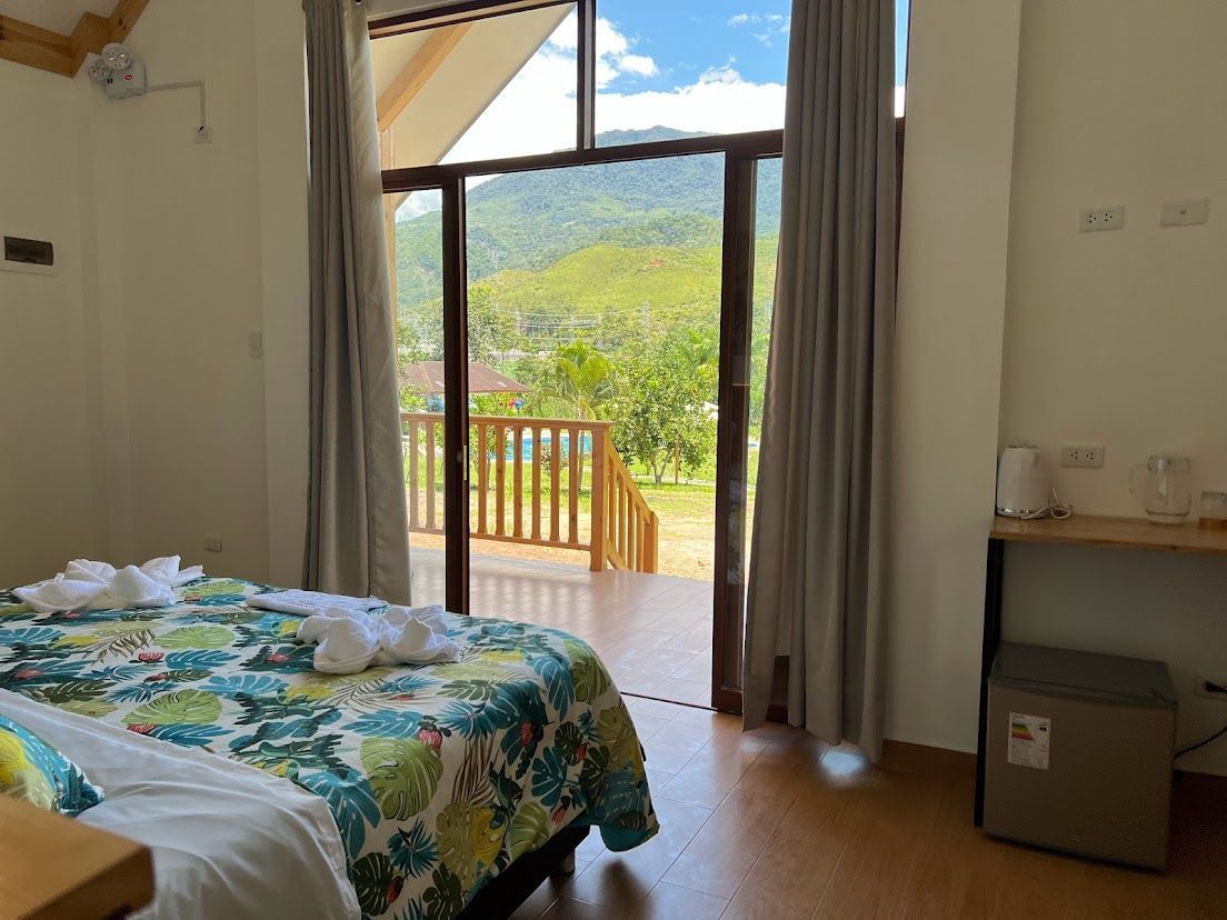 Habitación con colcha floral, puerta de balcón abierta y vista a la montaña más allá de las cortinas.