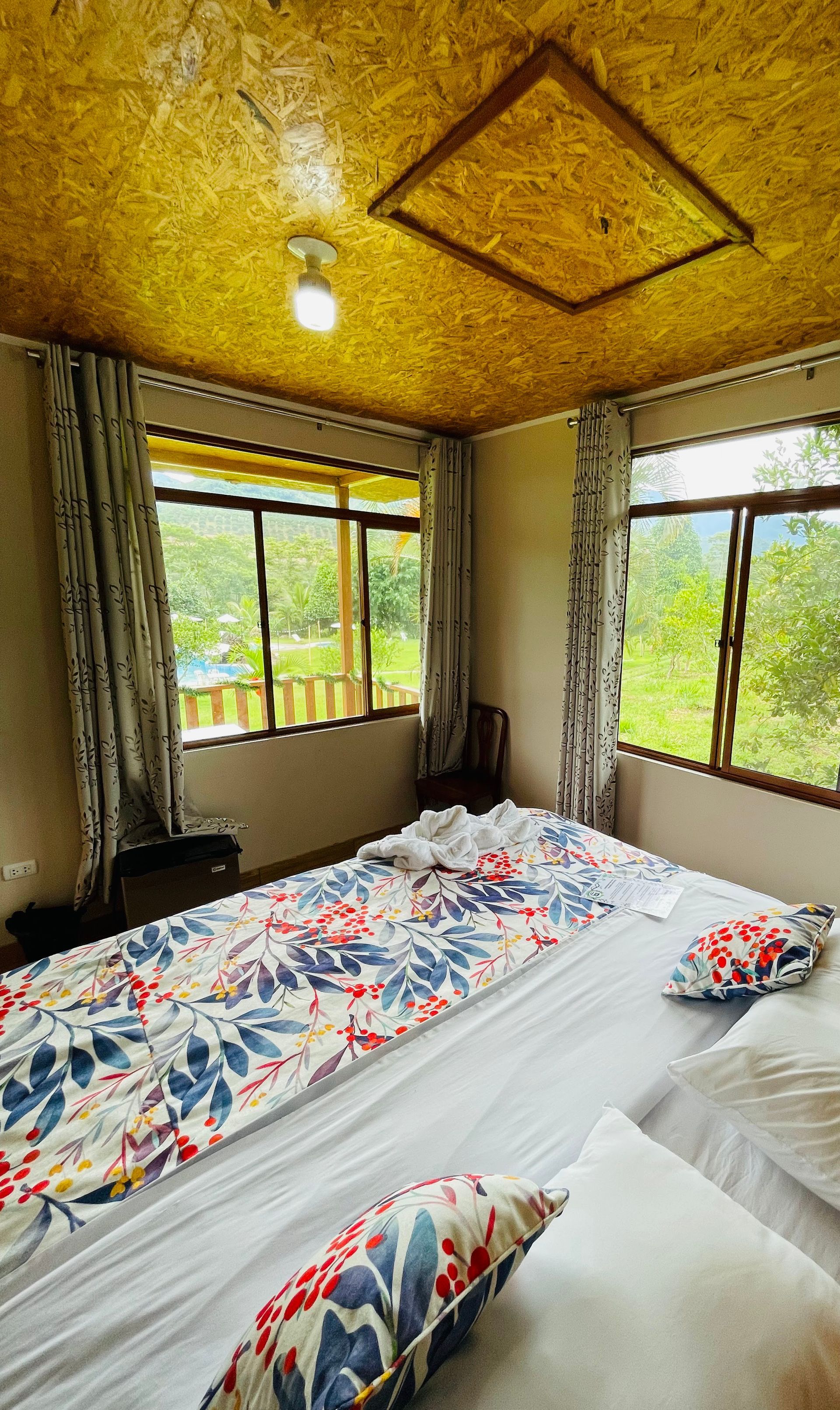 Habitación acogedora con cama, ropa de cama floral, grandes ventanales y techo amarillo texturizado.