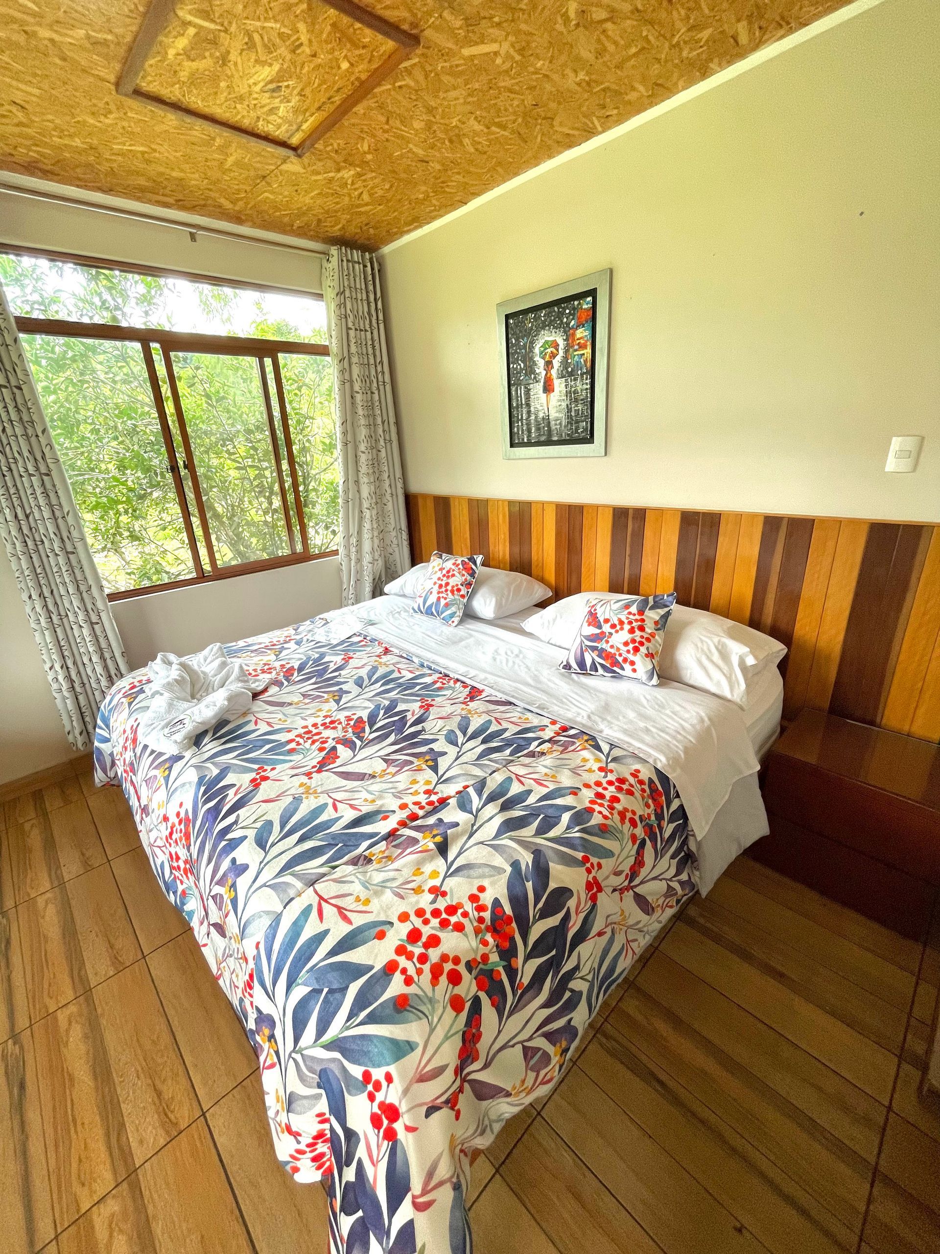 Dormitorio con colcha floral, suelo de madera y gran ventanal con cortinas transparentes.