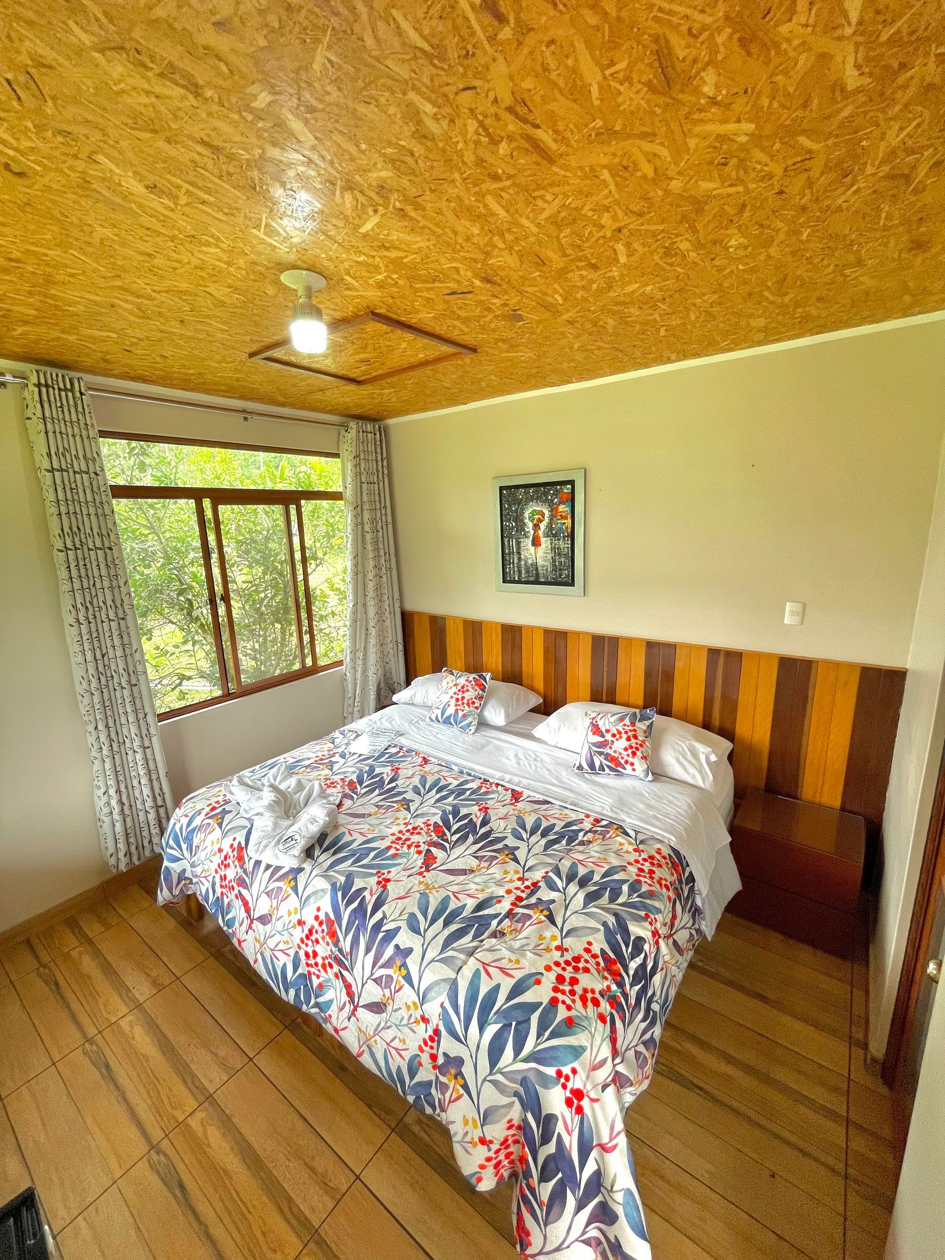 Acogedora habitación con colcha floral, suelo y techo de madera, cuadros enmarcados y ventana con cortinas.