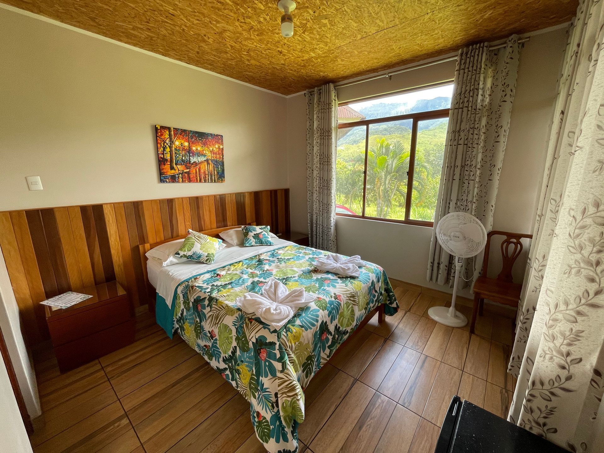 Habitación luminosa con una colcha estampada de flores, suelo de madera y luz natural que entra por una ventana con cortinas transparentes.