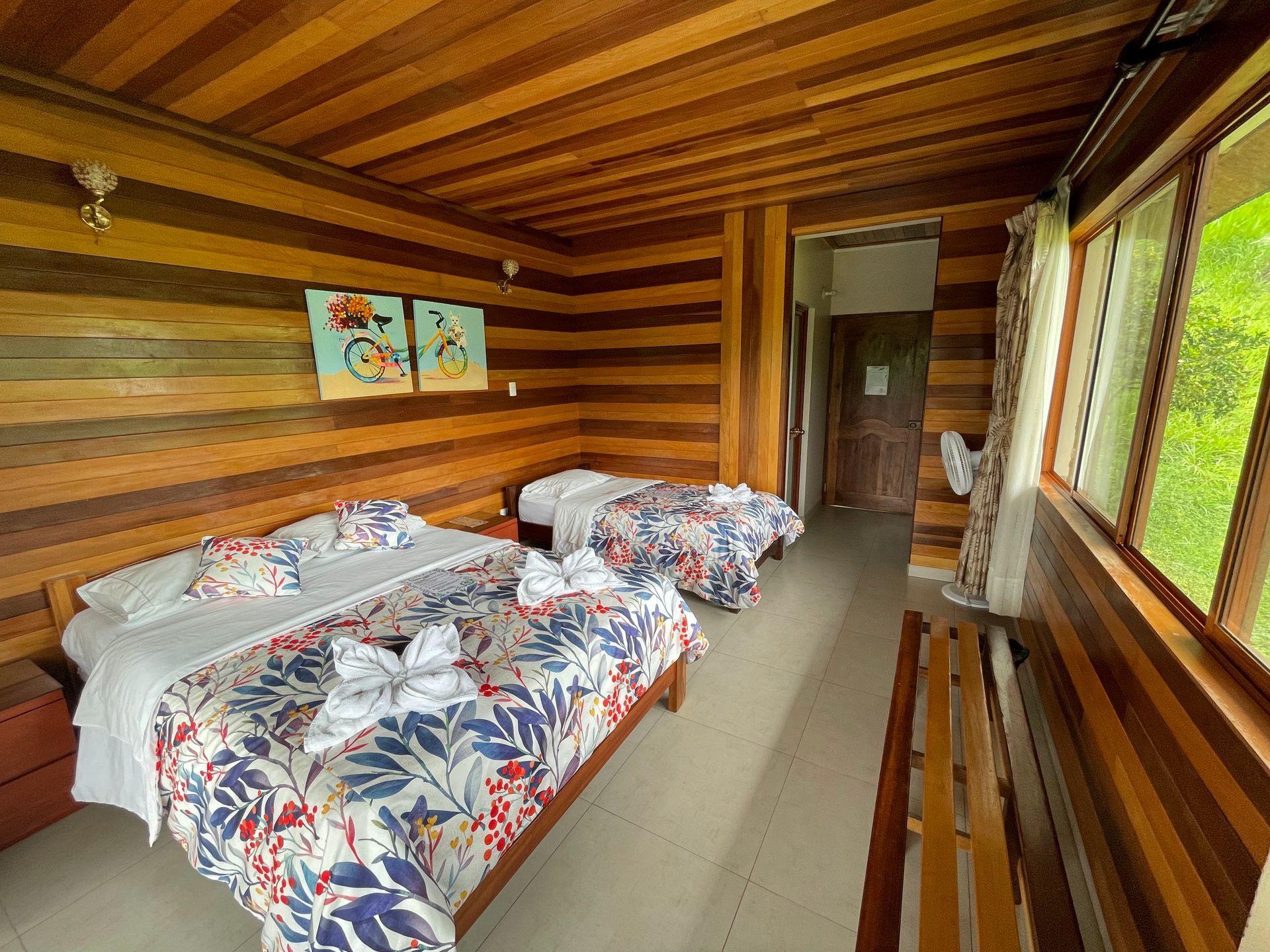 Dormitorio con paneles de madera, dos camas con motivos florales, una ventana y una puerta que da al pasillo.