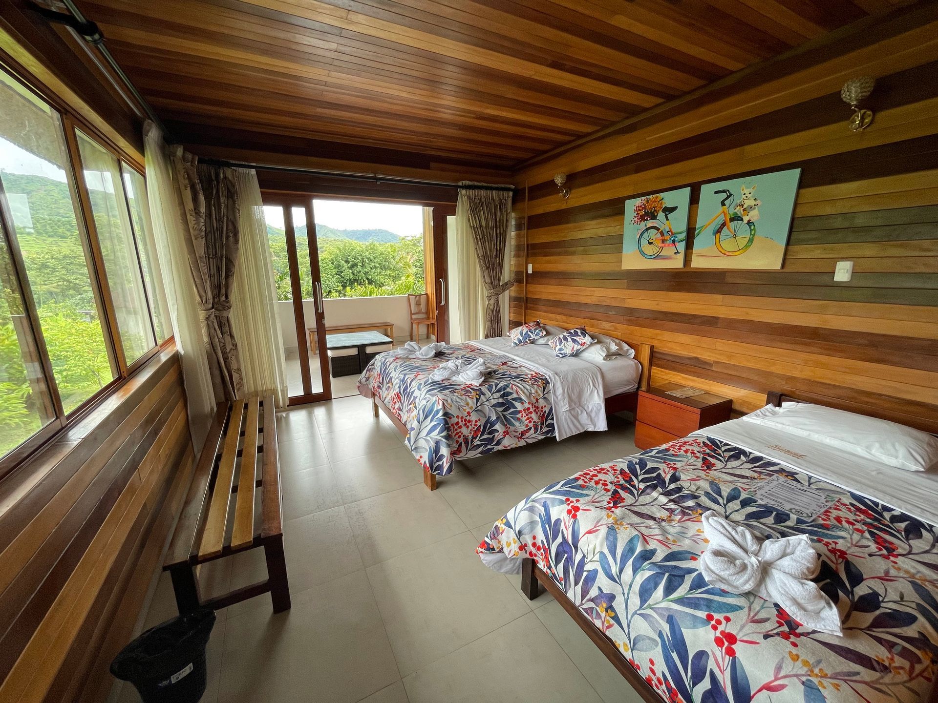 Habitación rústica con dos camas, paredes de madera, banco y balcón con vistas a la vegetación.