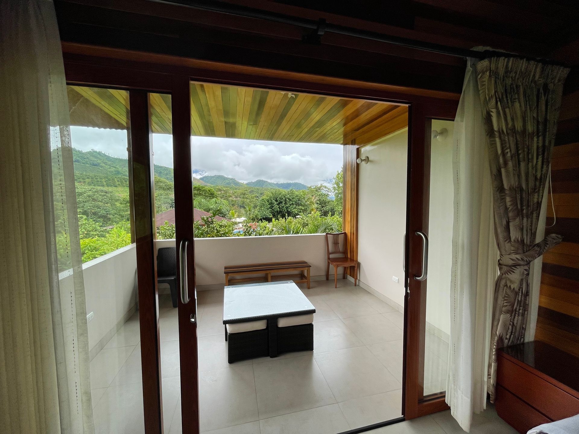 Balcón al aire libre con muebles de madera, cortinas blancas y una exuberante vista a la montaña verde.