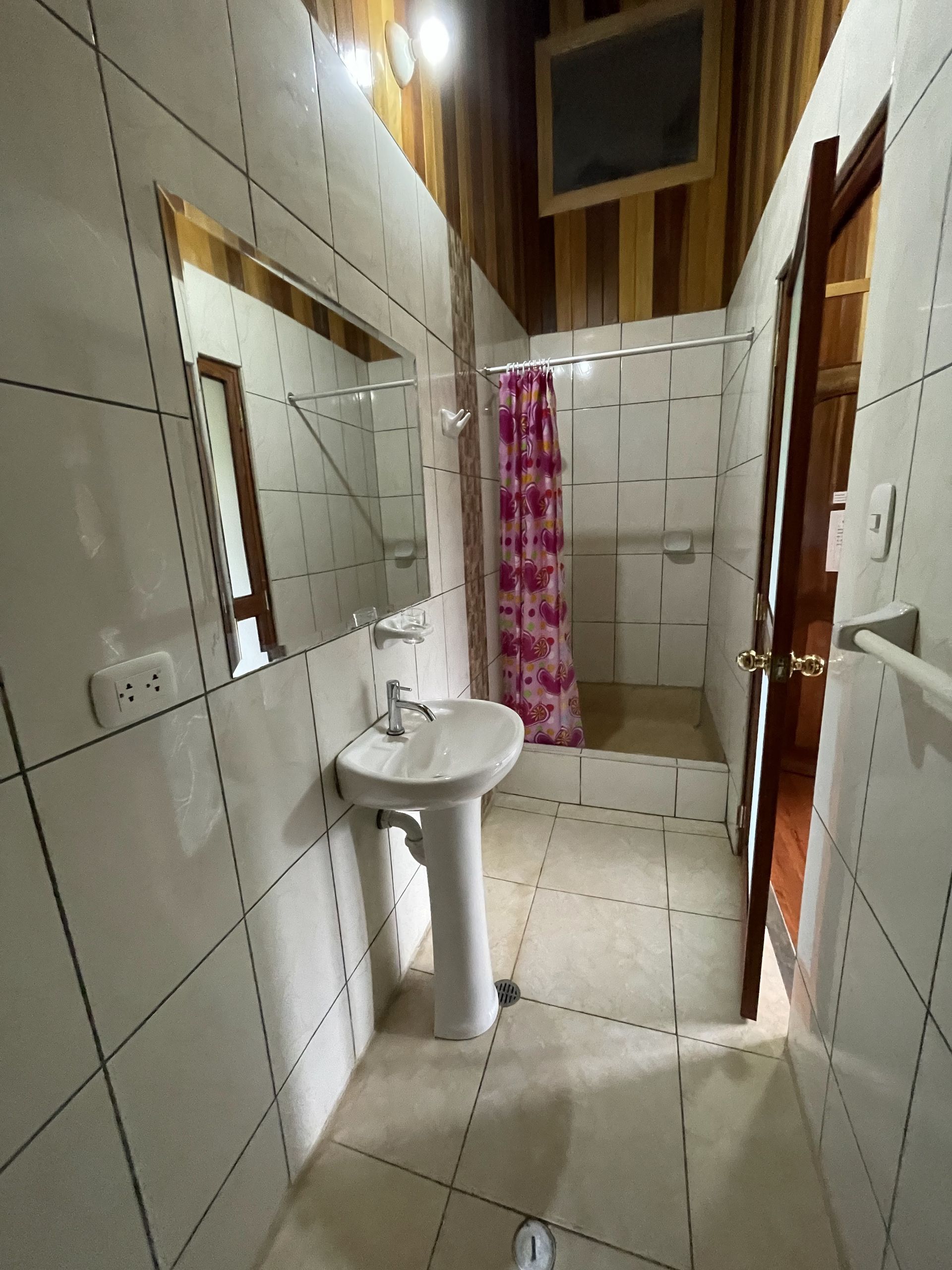 Baño pequeño con azulejos, lavabo, espejo, puerta abierta y cortina de ducha rosa.