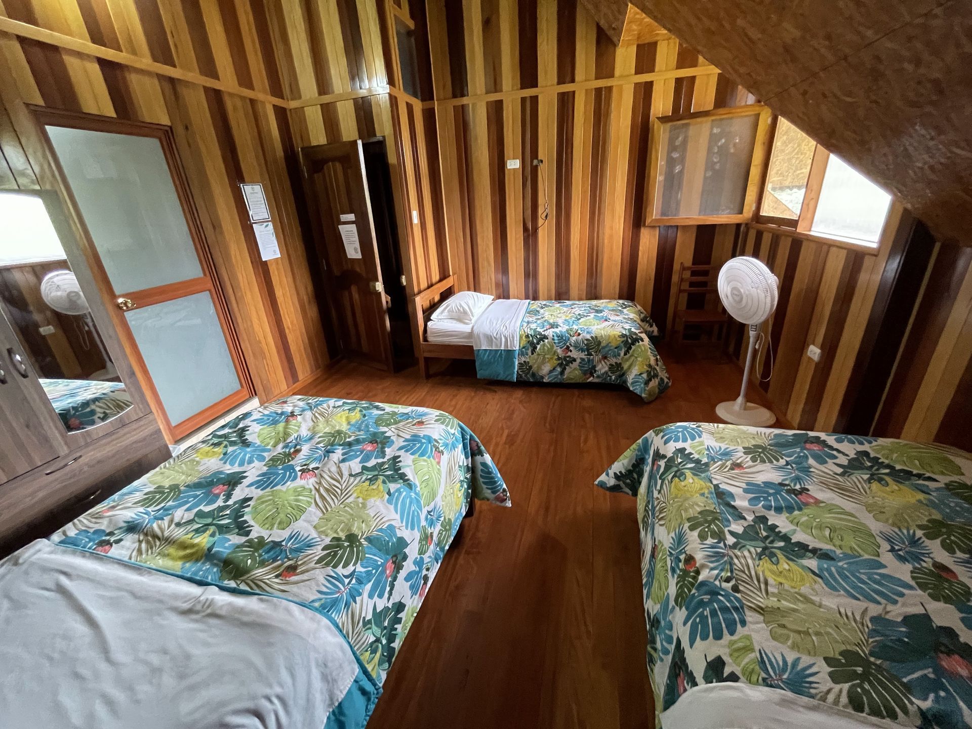 Pequeña habitación abuhardillada con tres camas, paredes revestidas de madera y luz natural cálida que entra por una ventana.