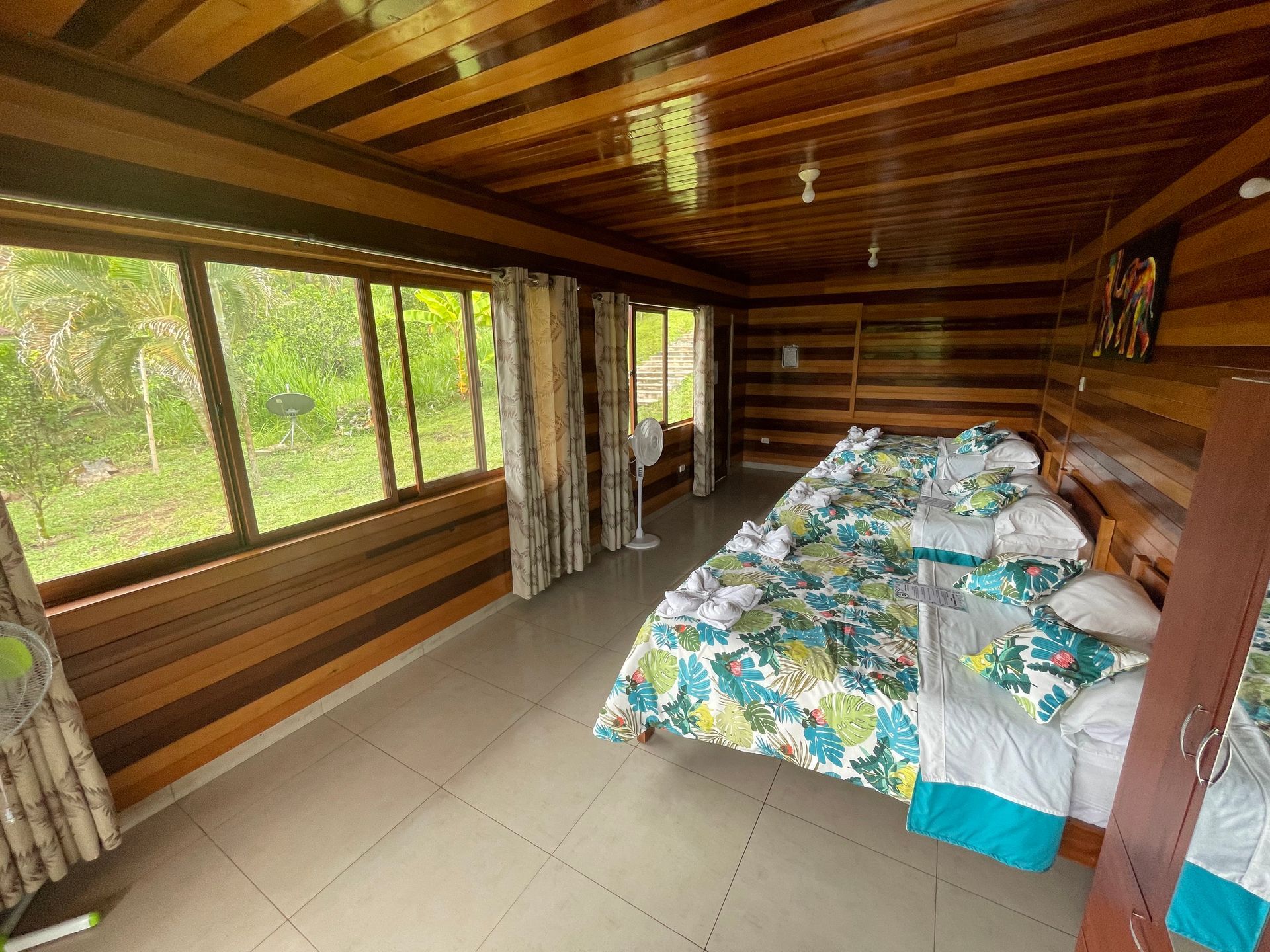 Habitación en una cabaña de madera con cama, grandes ventanas y cortinas con vistas a árboles verdes.