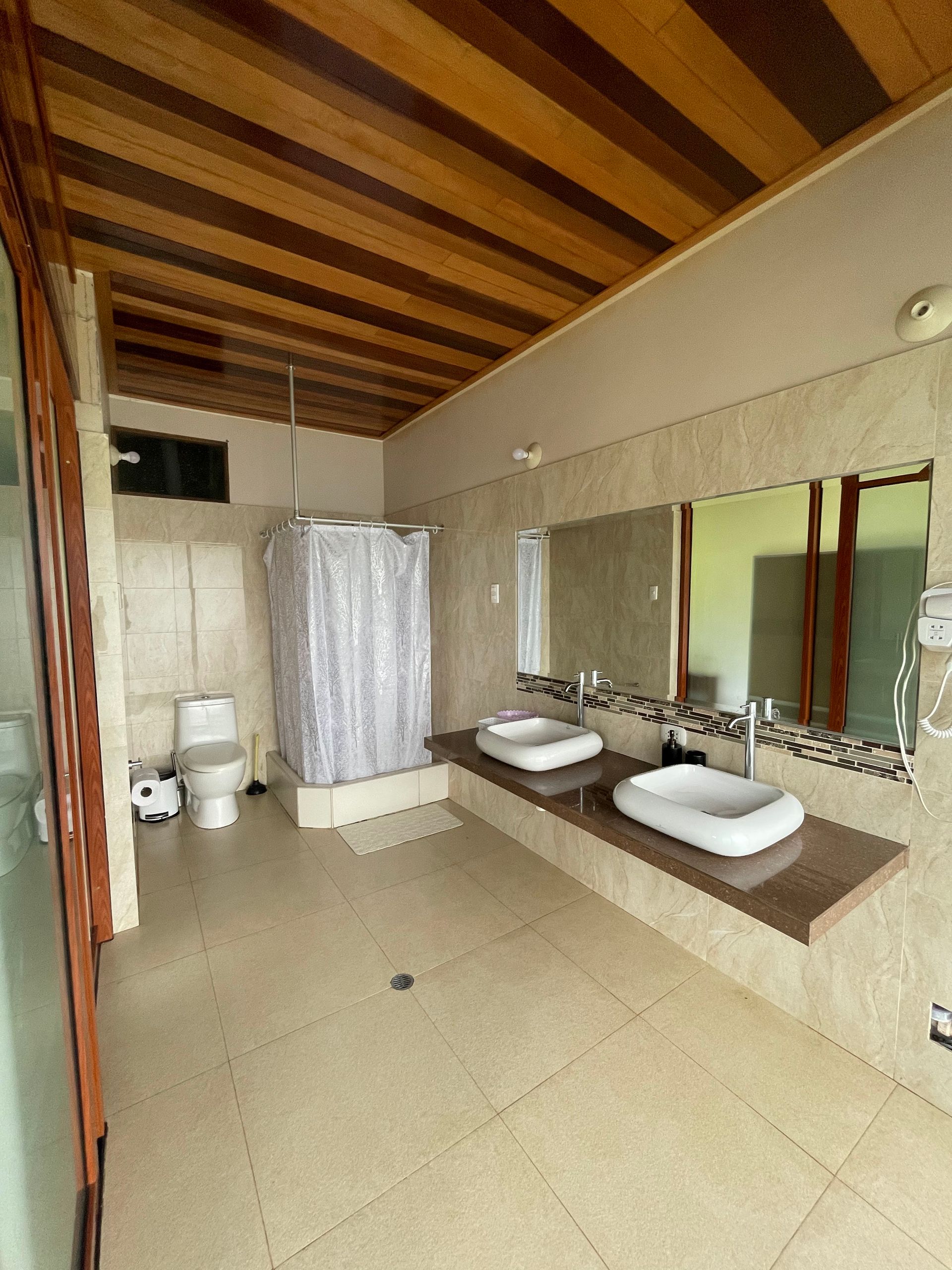 Baño moderno con dos lavabos, espejos, inodoro, cortina de ducha y techo de listones de madera.