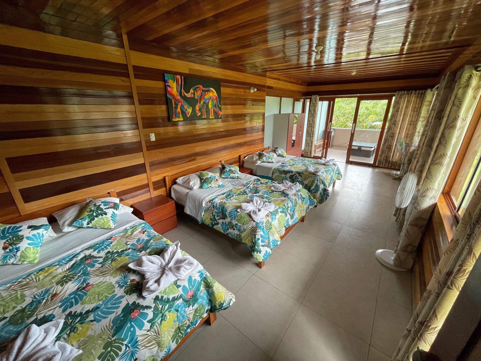 Habitación de madera con tres camas con estampado tropical, suelo de baldosas y puertas que dan a un balcón luminoso.