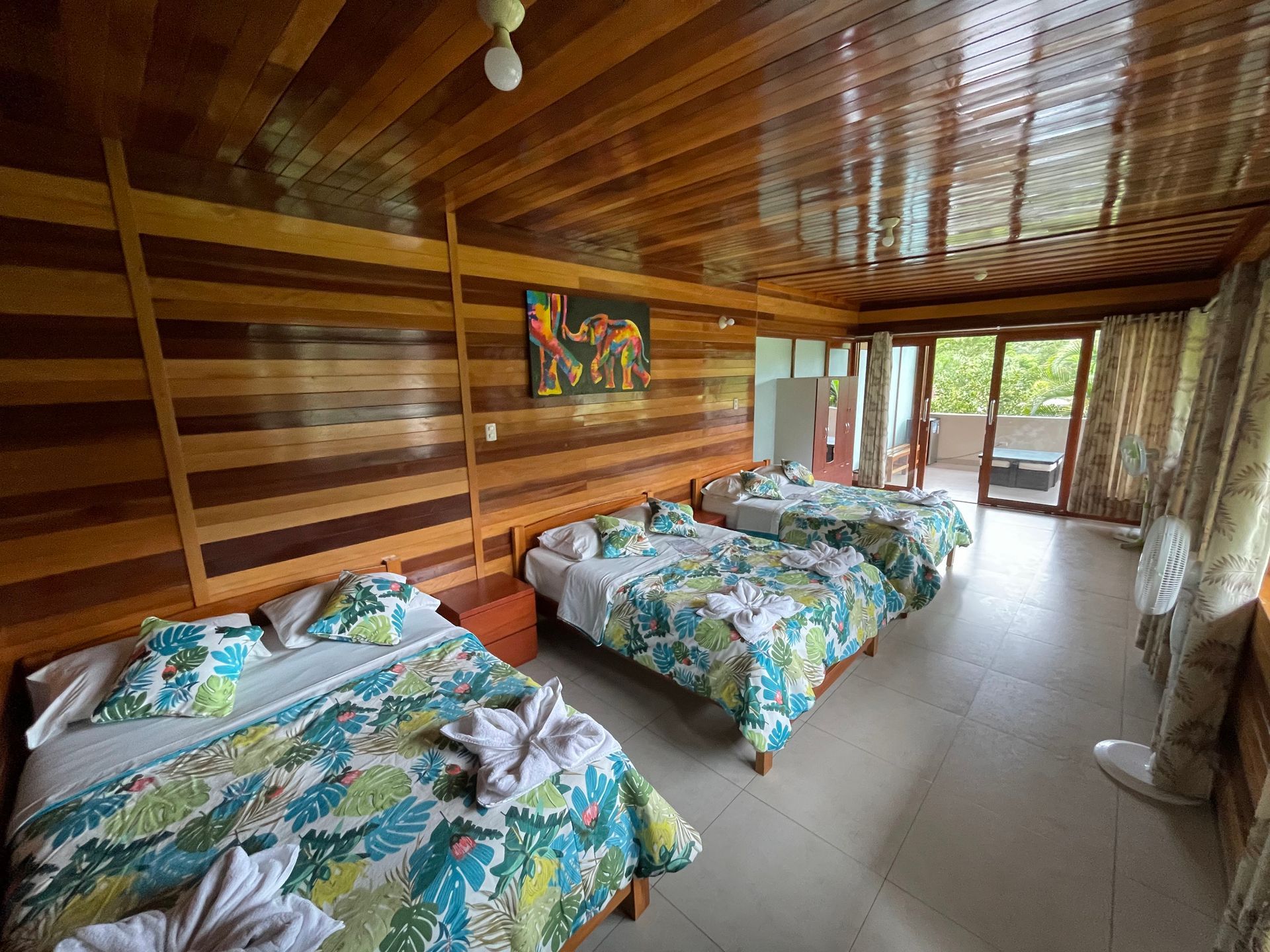 Dormitorio con paneles de madera, dos camas con estampado tropical y vistas a una exuberante vegetación a través de puertas corredizas de cristal.