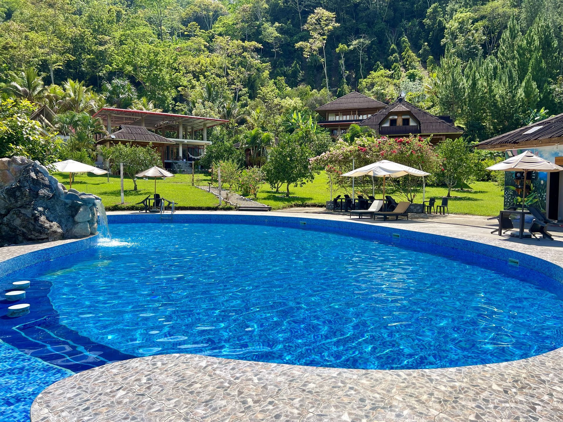 Piscina del complejo turístico con agua azul brillante, tumbonas, sombrillas y una exuberante ladera verde al fondo.