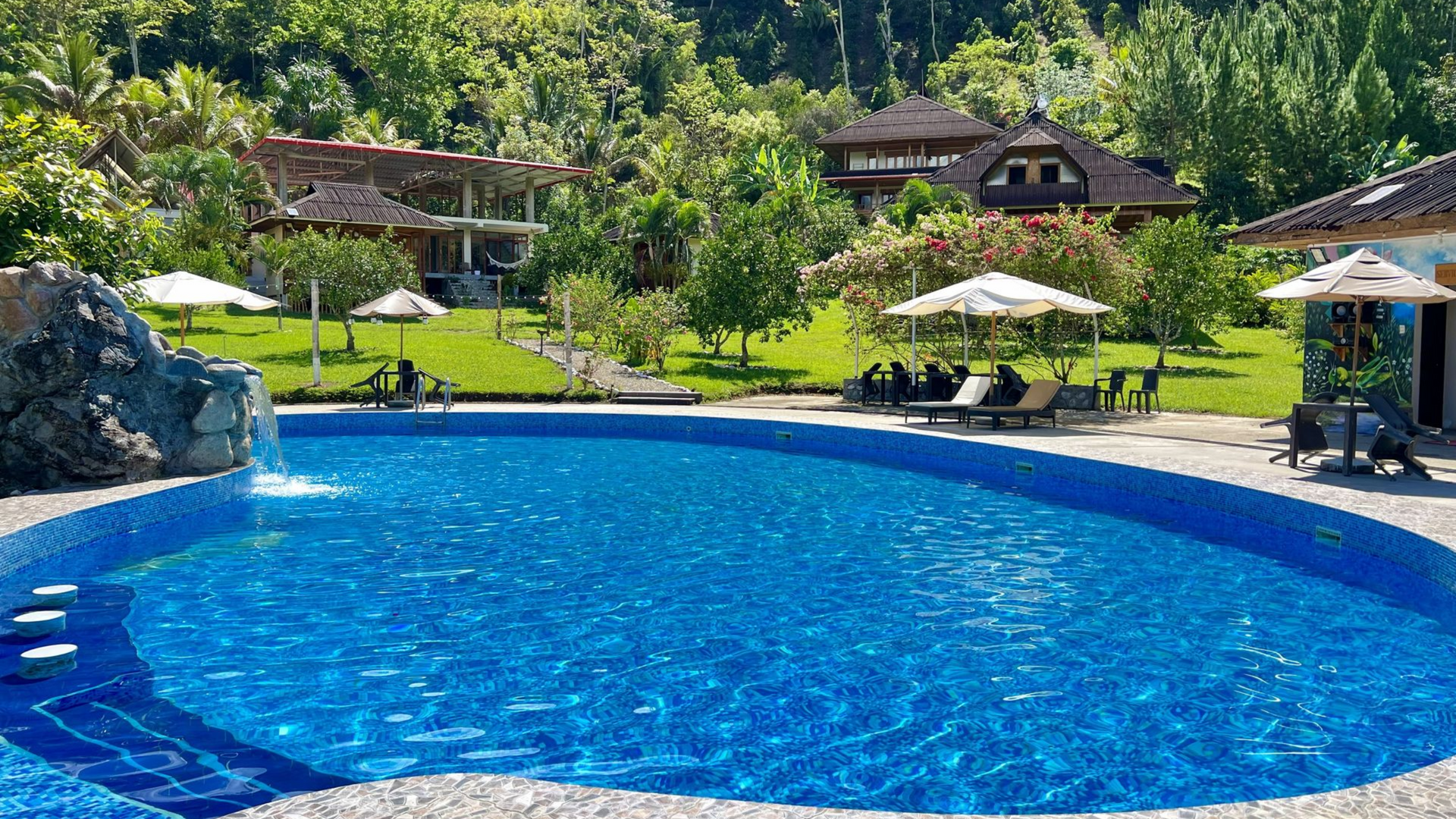 Piscina azul de un complejo turístico con tumbonas, sombrillas y exuberante vegetación en un día soleado.