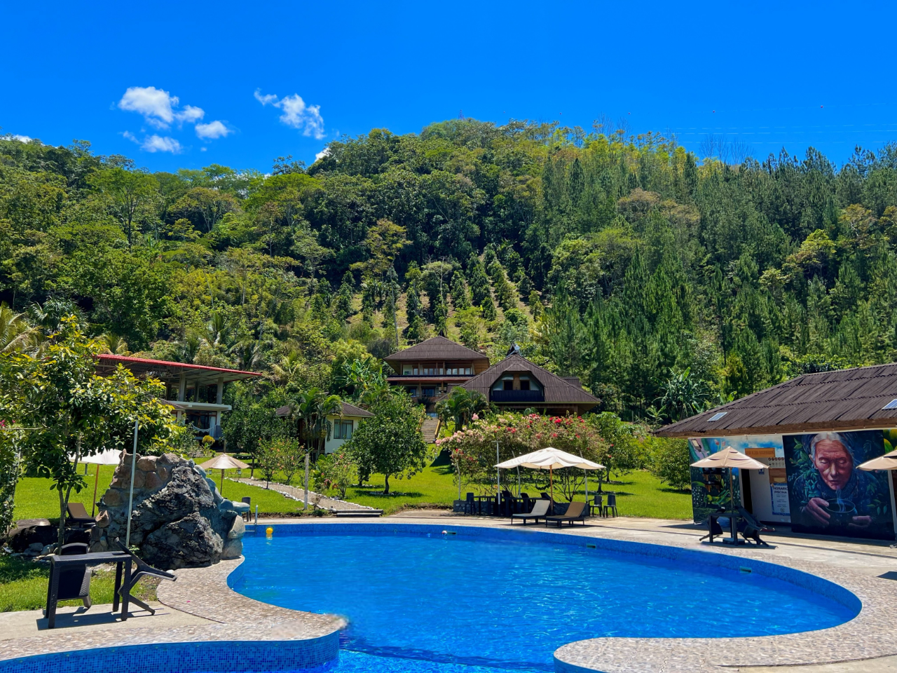 Piscina del complejo turístico con tumbonas y sombrillas, con vistas a verdes colinas bajo un cielo azul brillante.