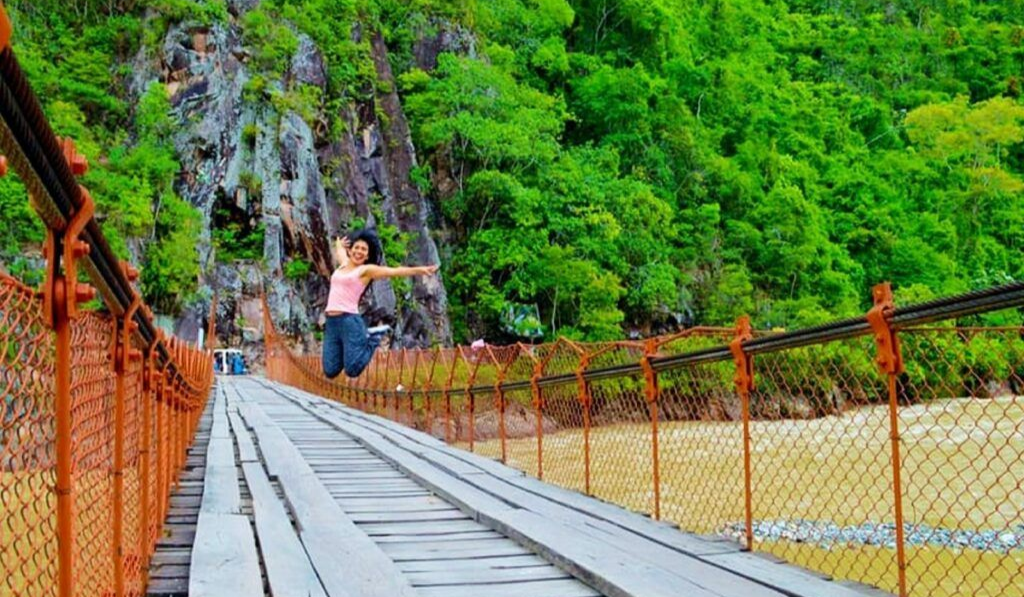 Mujer cruzando un puente colgante de madera en un entorno tropical exuberante y verde.