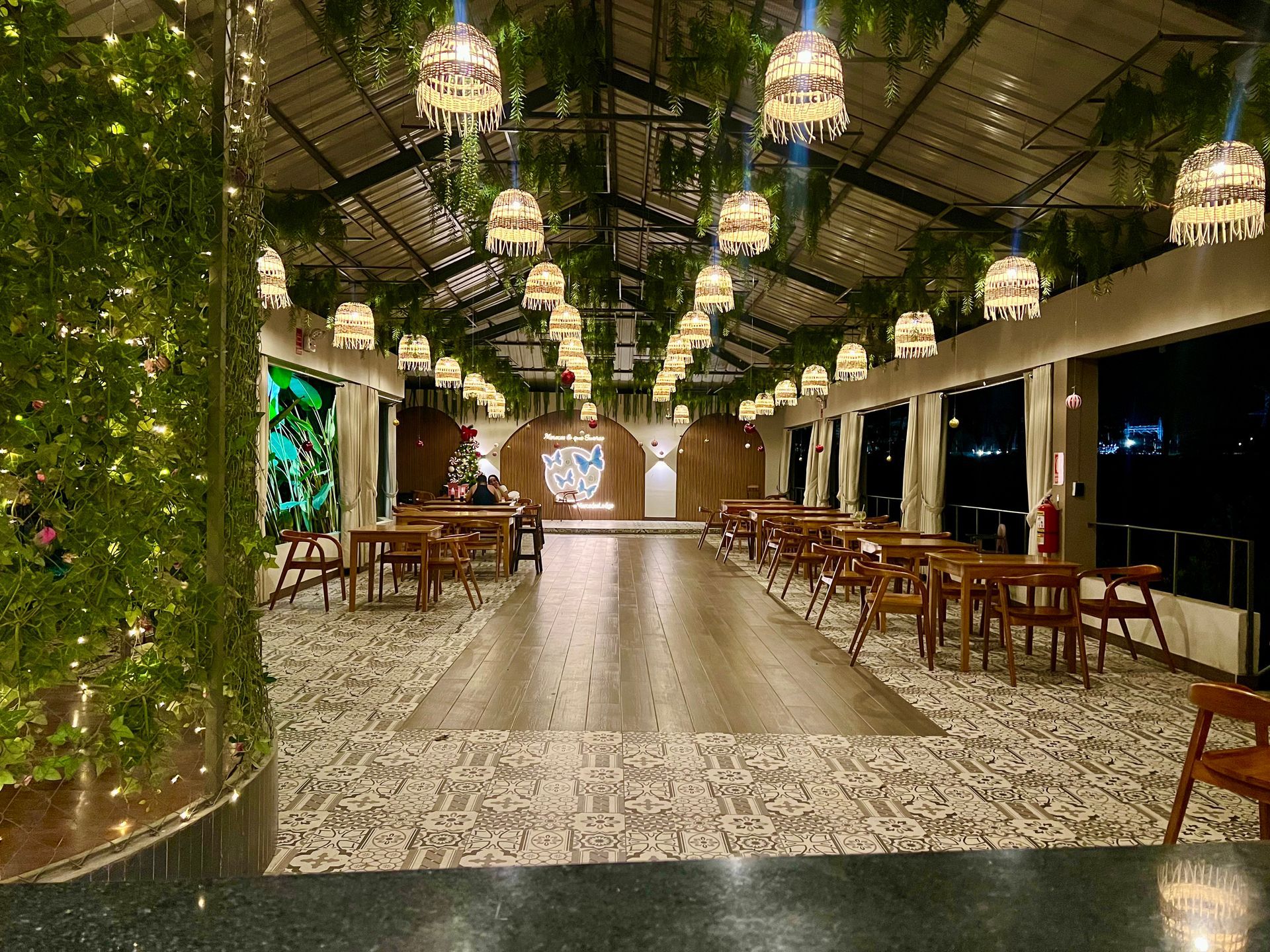 Comedor exterior cubierto con faroles colgantes, mesas de madera y suelo de baldosas estampadas por la noche.